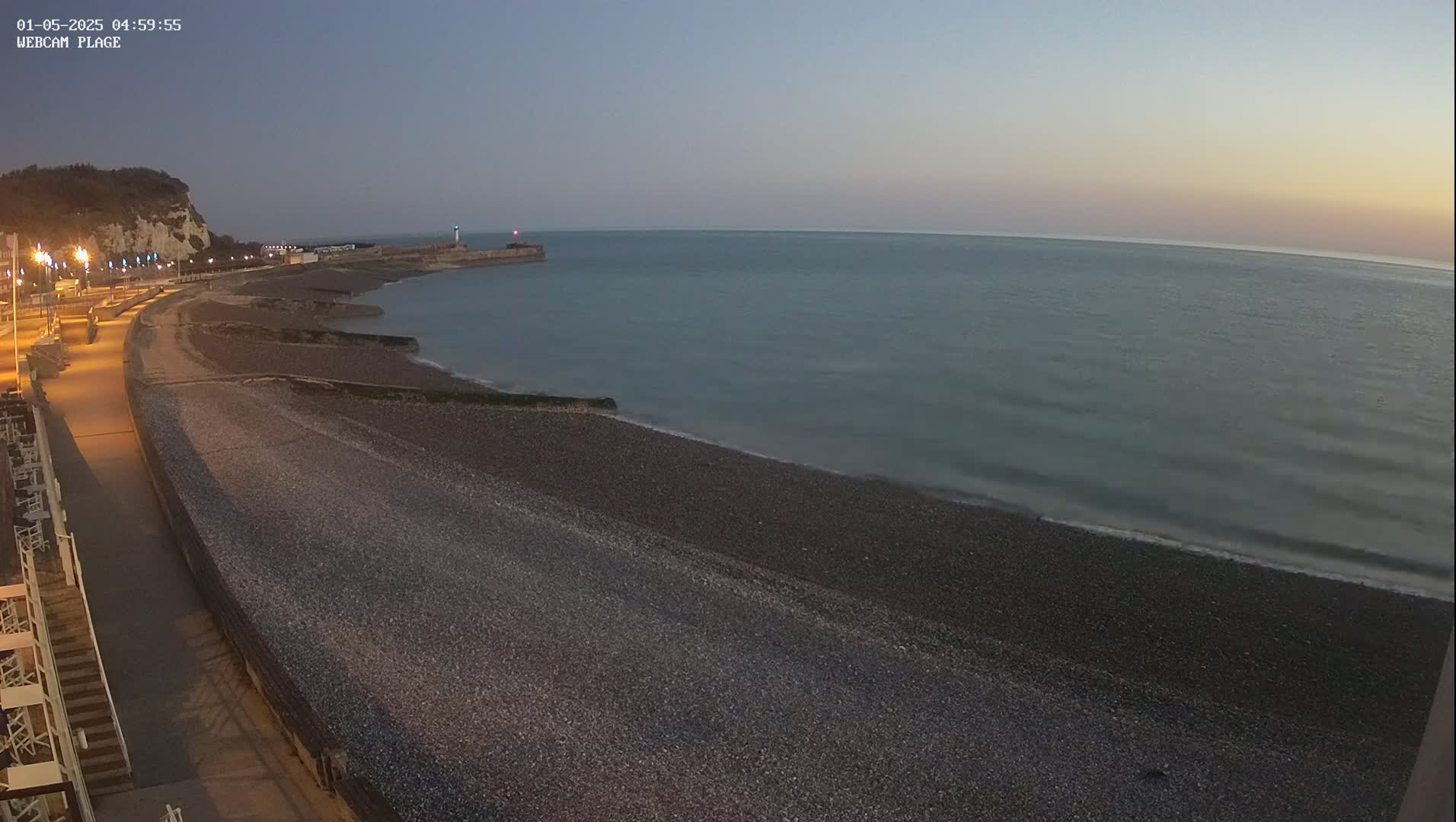 A dark pebble beach and calm sea are visible at twilight, with a paved walkway and distant cliffs under a clear sky.
