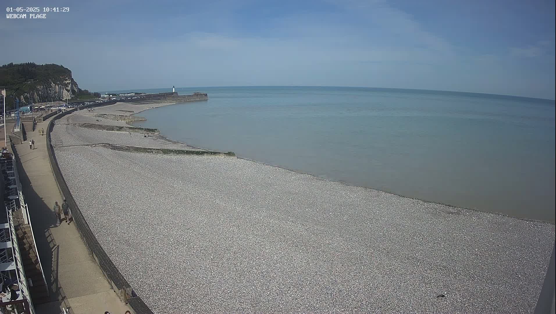A pebble beach stretches along a calm sea under a clear blue sky, with a walkway and a few people visible.