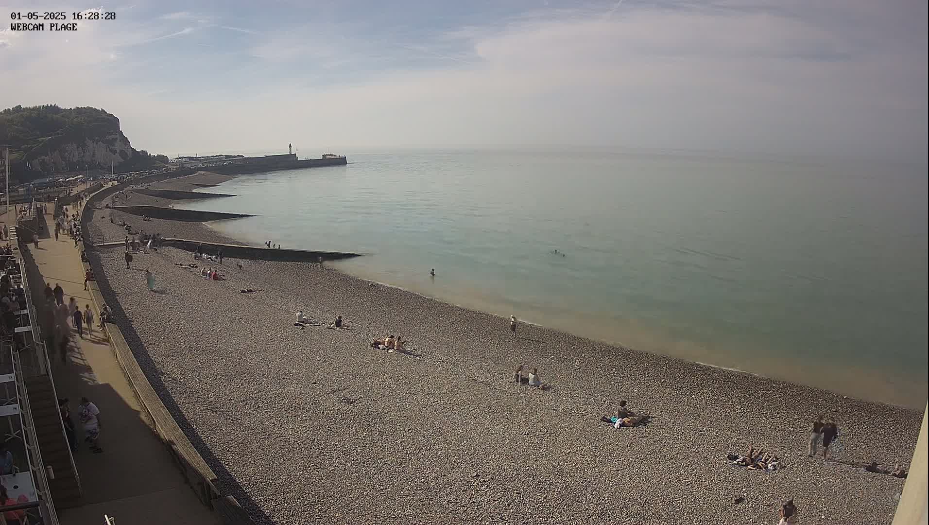 A pebble beach with people sunbathing and walking along a promenade, next to calm, light-green sea under a mostly sunny sky.