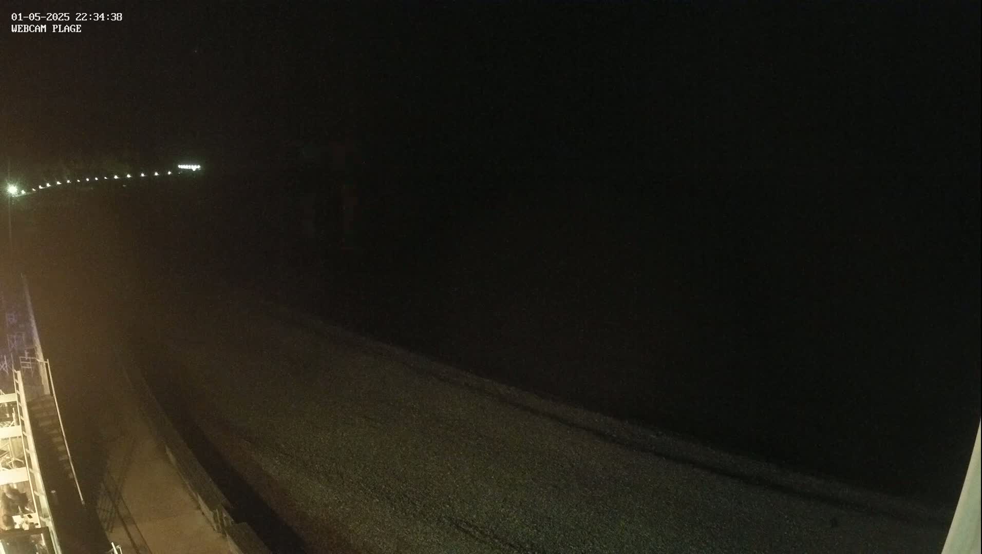 A dark, nighttime view of a pebble beach next to a dark body of water, illuminated only by faint lights in the distance.