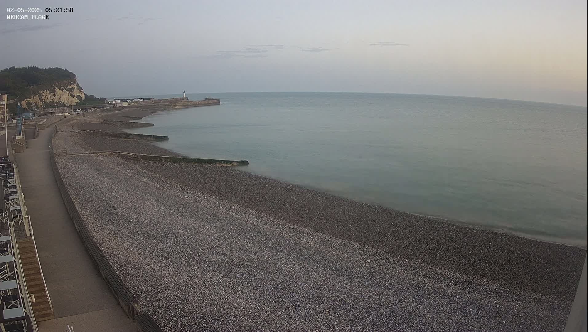 A pebble beach stretches along a calm sea under a pale, overcast sky, with a walkway and cliff visible in the background.