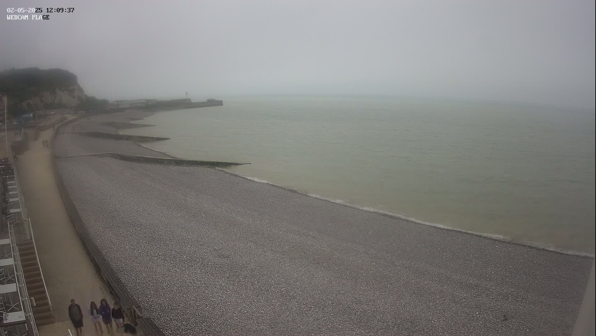 A foggy day at a pebble beach shows a calm sea, a walkway, and a few people walking near a cliffside.