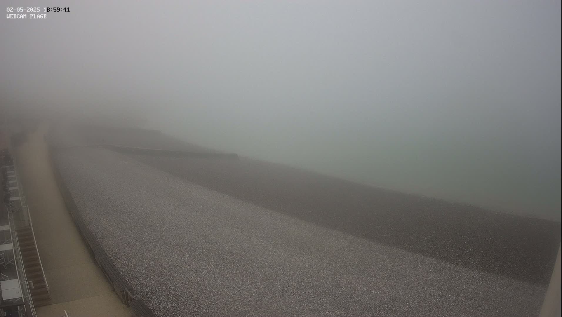 A foggy view shows a pebble beach and a walkway next to a building.
