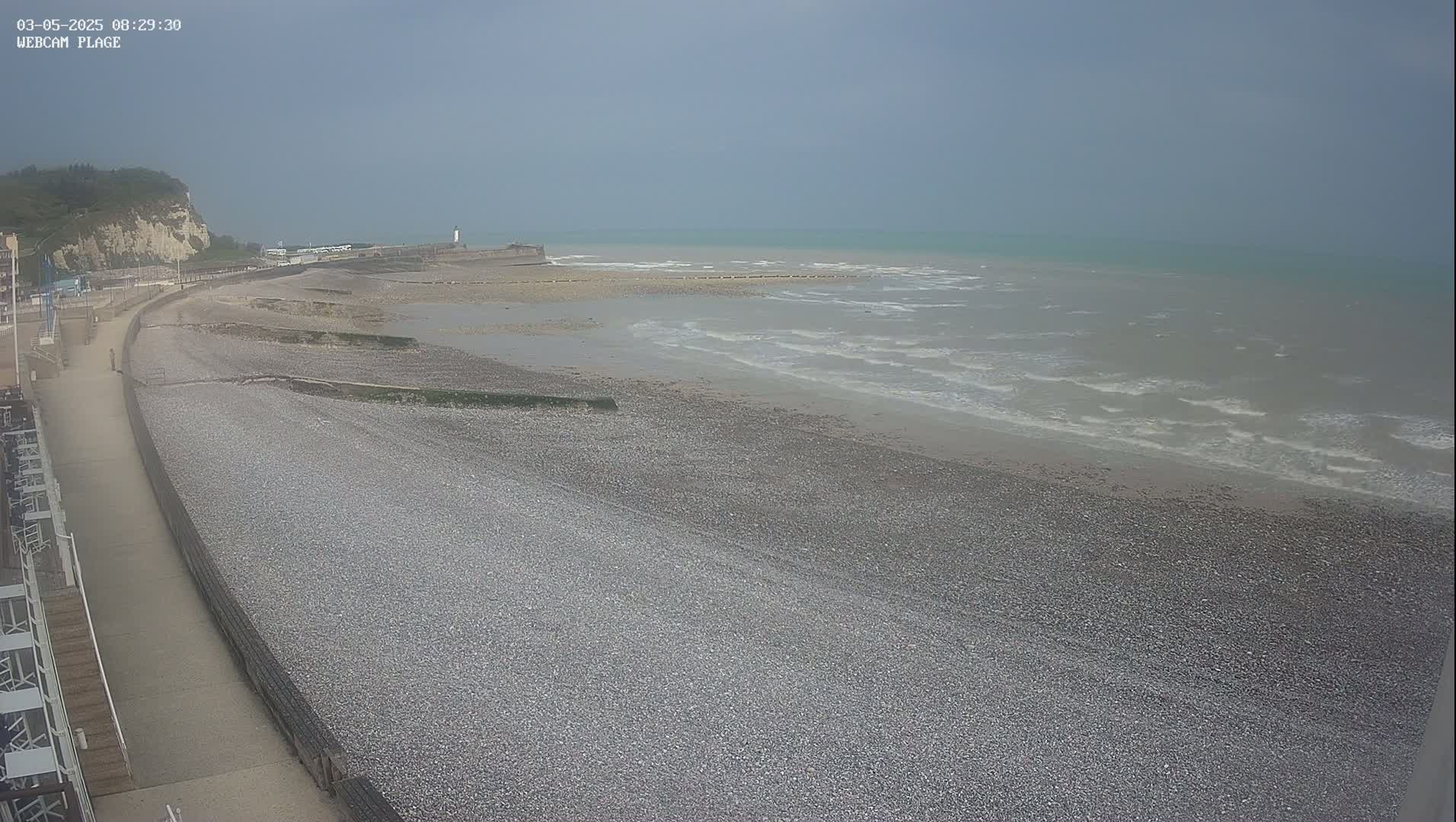 A pebble beach stretches alongside a paved walkway, meeting a calm, gray sea under an overcast sky.