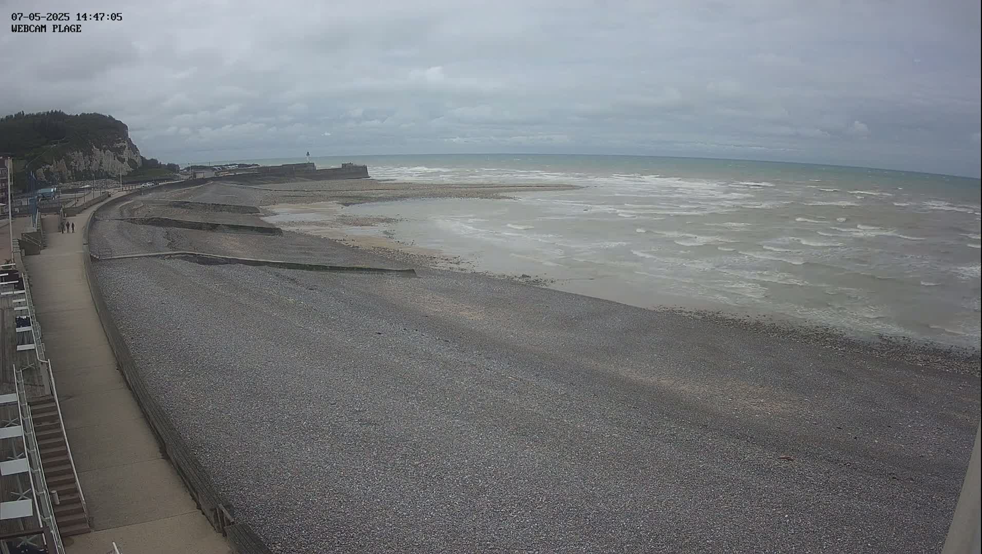 A pebble beach with a walkway and a few people walking stretches alongside a calm sea under an overcast sky.