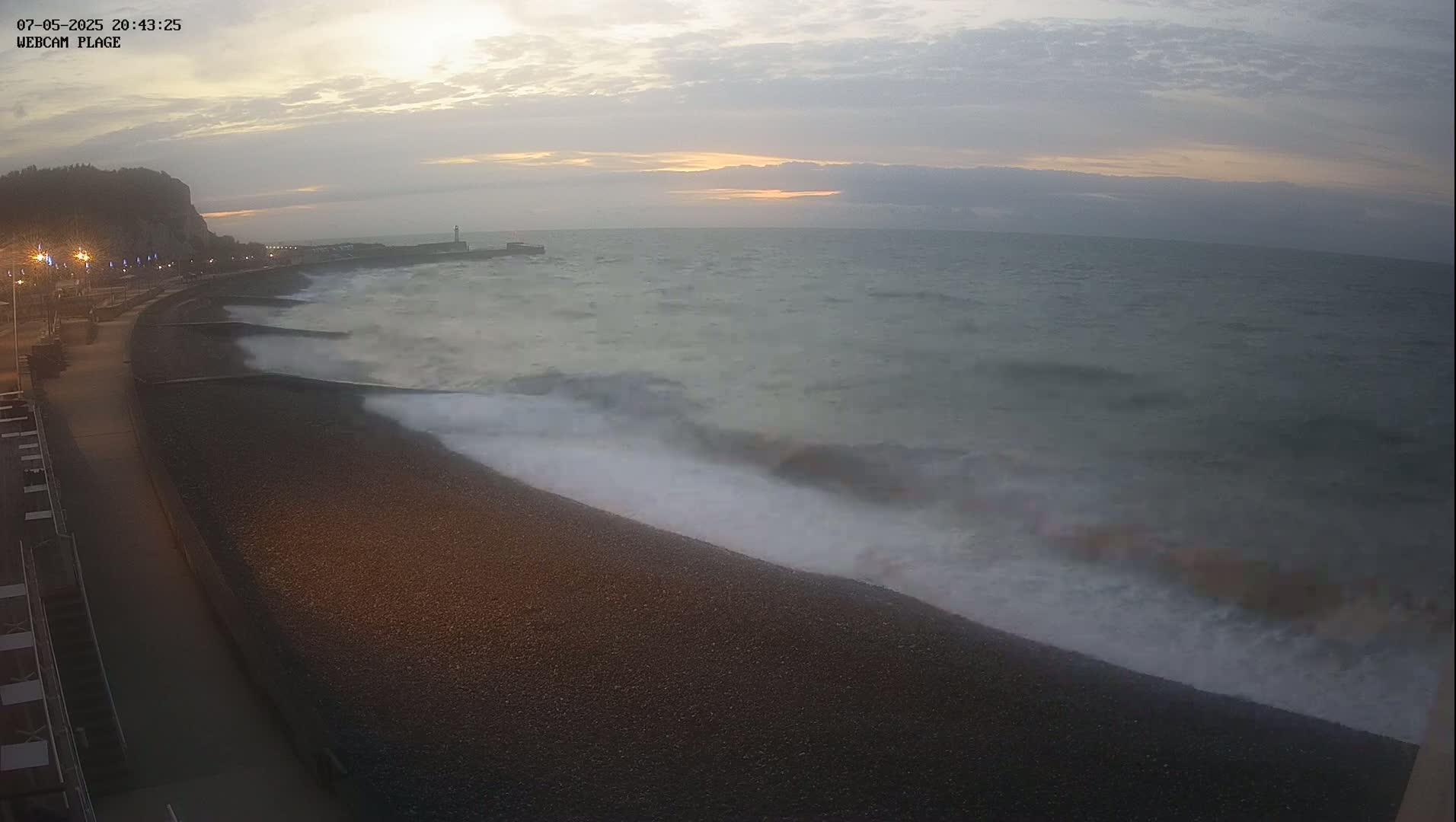 Waves crash on a dark pebble beach at dusk under a cloudy sky.
