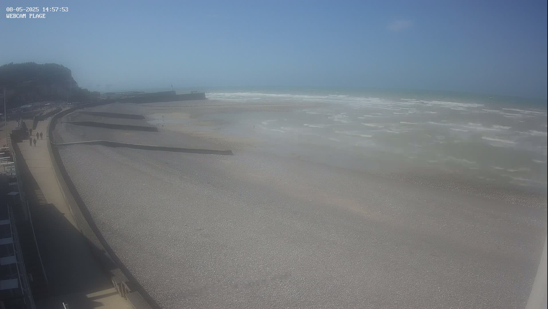 A pebble beach with a sea wall and a paved walkway extends to a hazy, windy ocean under a mostly clear sky.