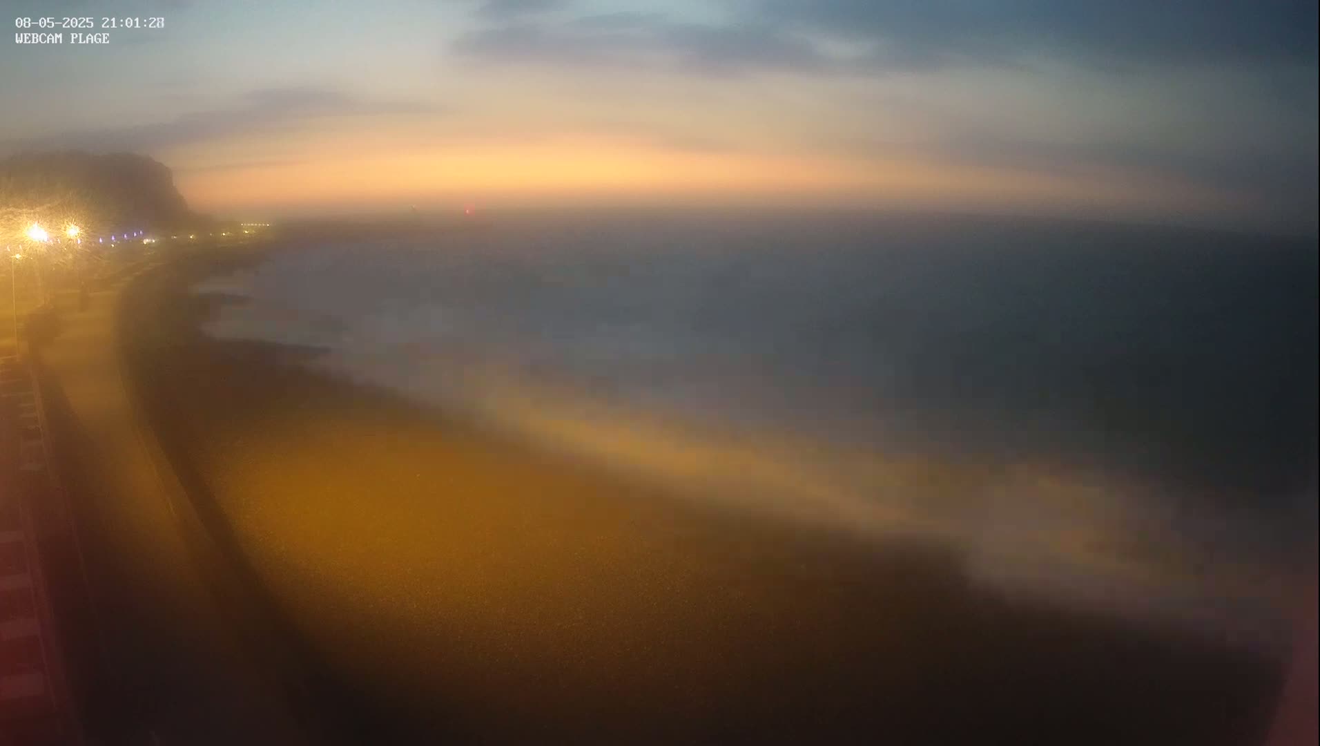 A hazy, orange-tinged sunset illuminates a dark beach and calm ocean, with a dimly lit roadway visible in the foreground.