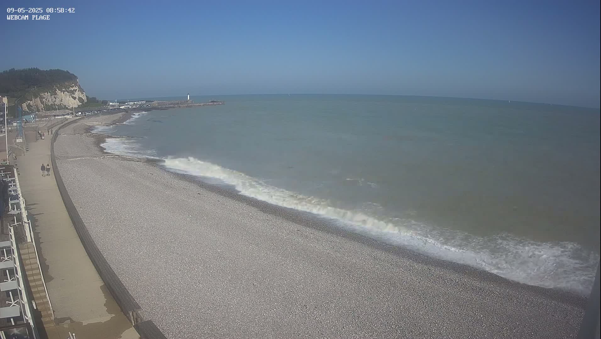 A pebble beach with gentle waves lapping the shore on a clear, sunny day, with a paved walkway and distant cliffs visible.