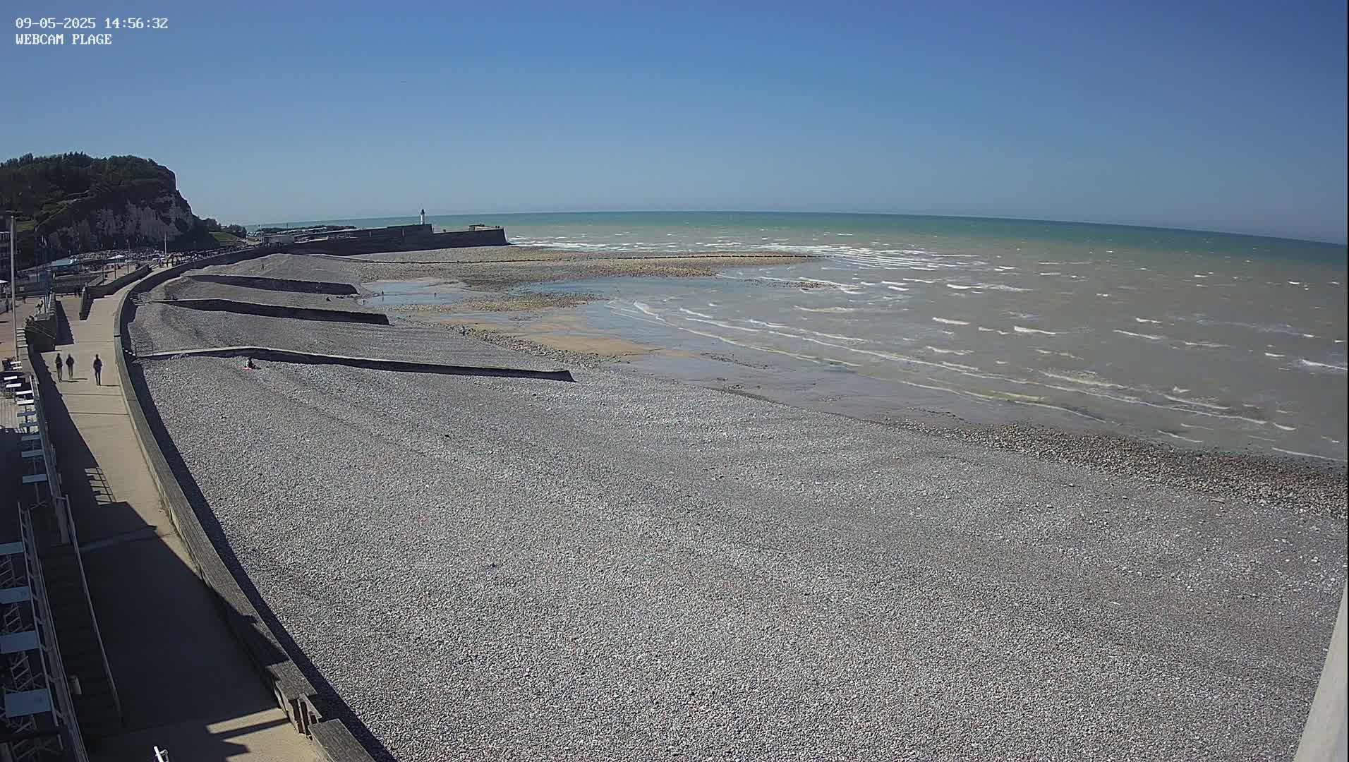 A pebble beach stretches along a concrete promenade under a clear blue sky, with small waves lapping the shore.