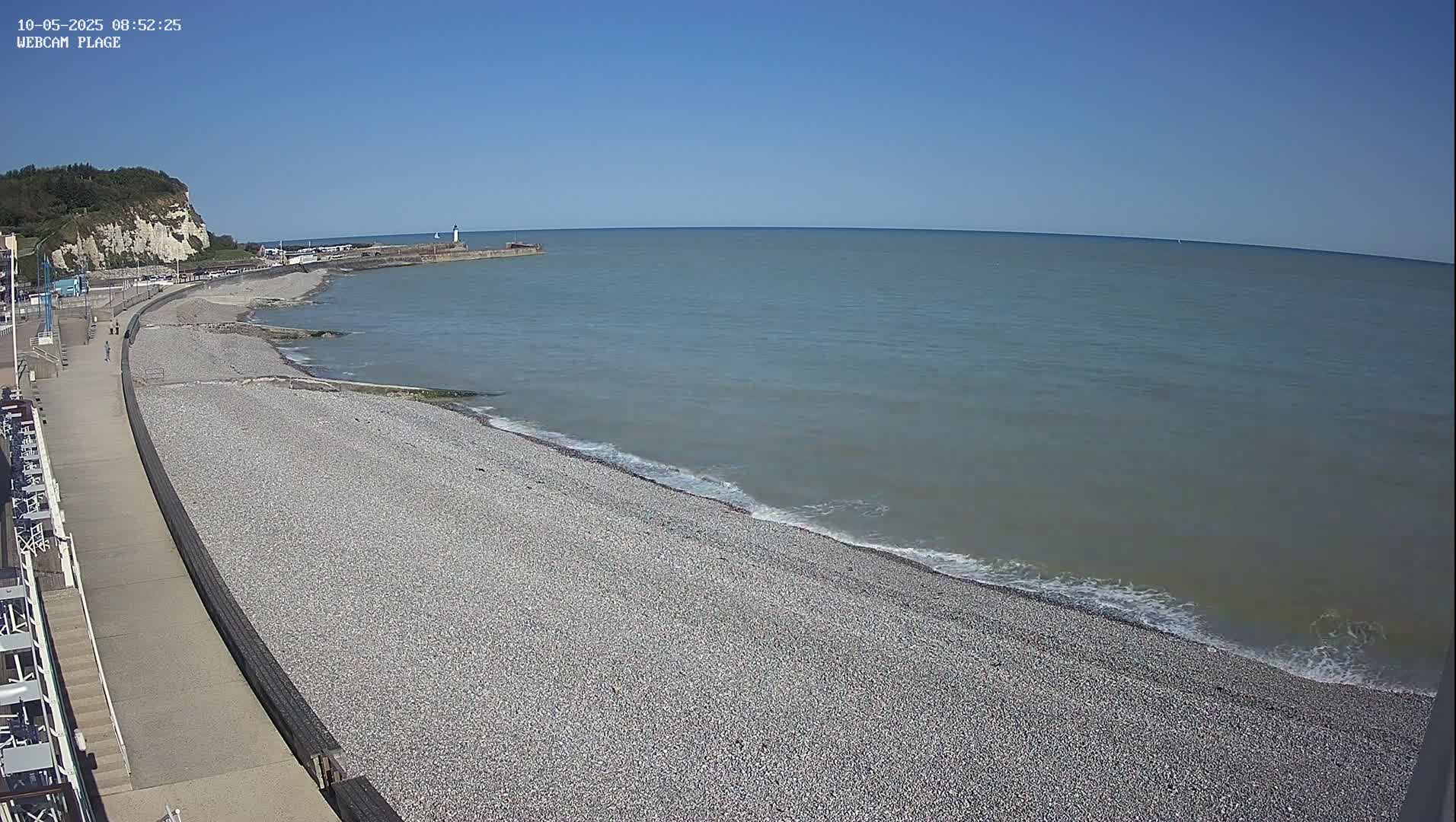 A pebble beach stretches along a calm, grey-blue sea under a clear blue sky, with a paved walkway and white buildings bordering the beach.