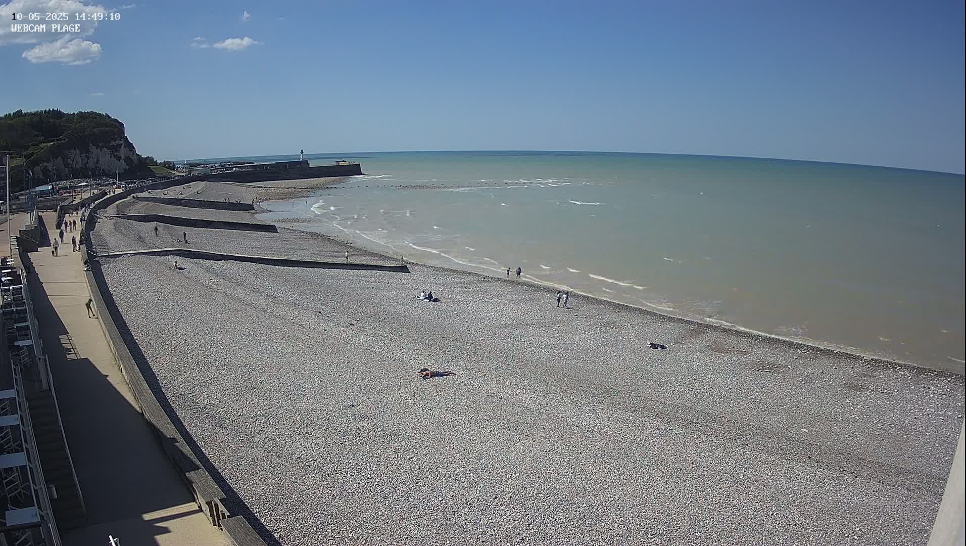 A pebble beach with scattered people on a sunny day next to a calm sea with small waves, backed by a cliff and promenade.