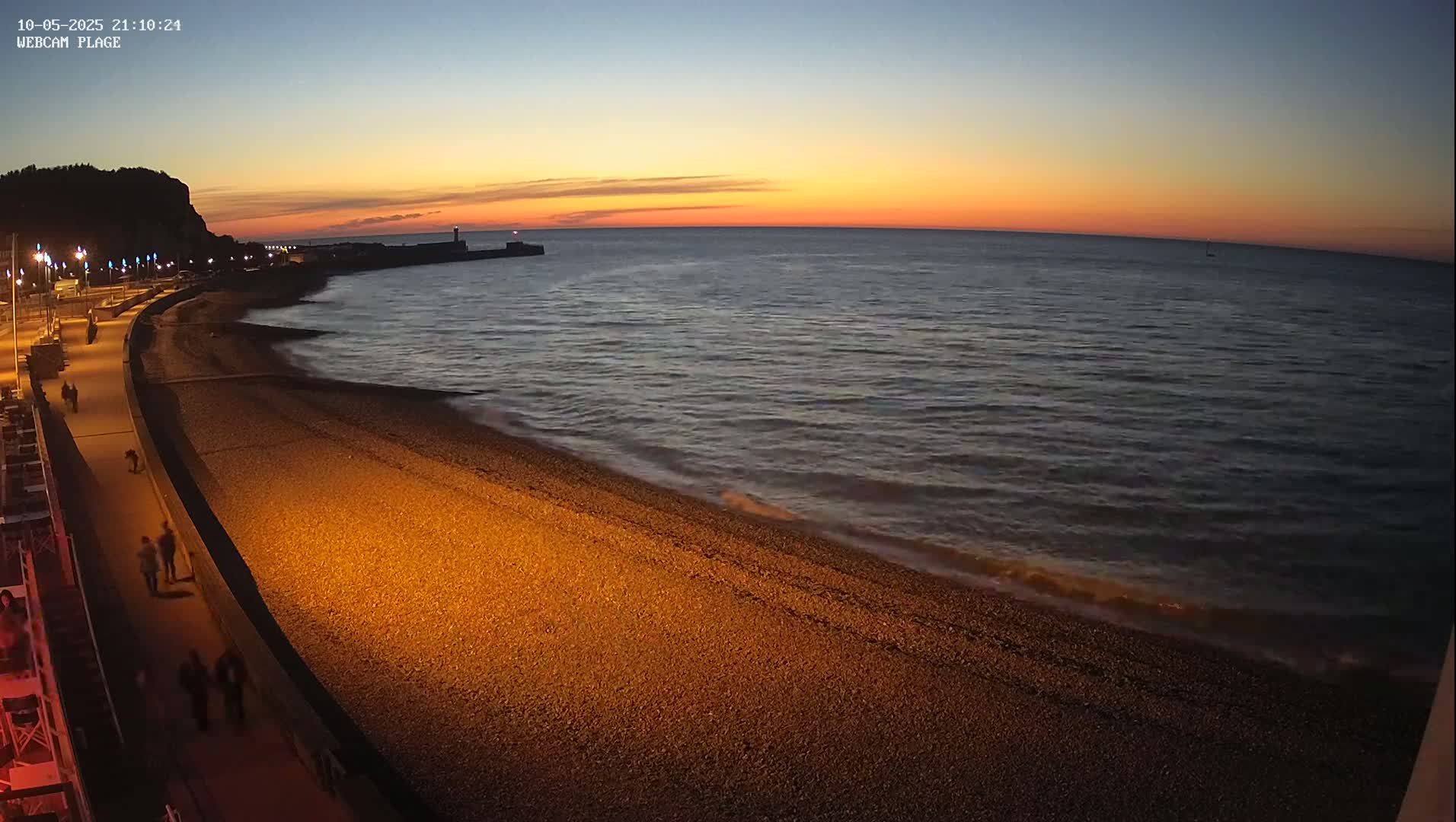 A pebble beach and illuminated promenade are shown at dusk, with gentle waves lapping the shore under a sunset sky.