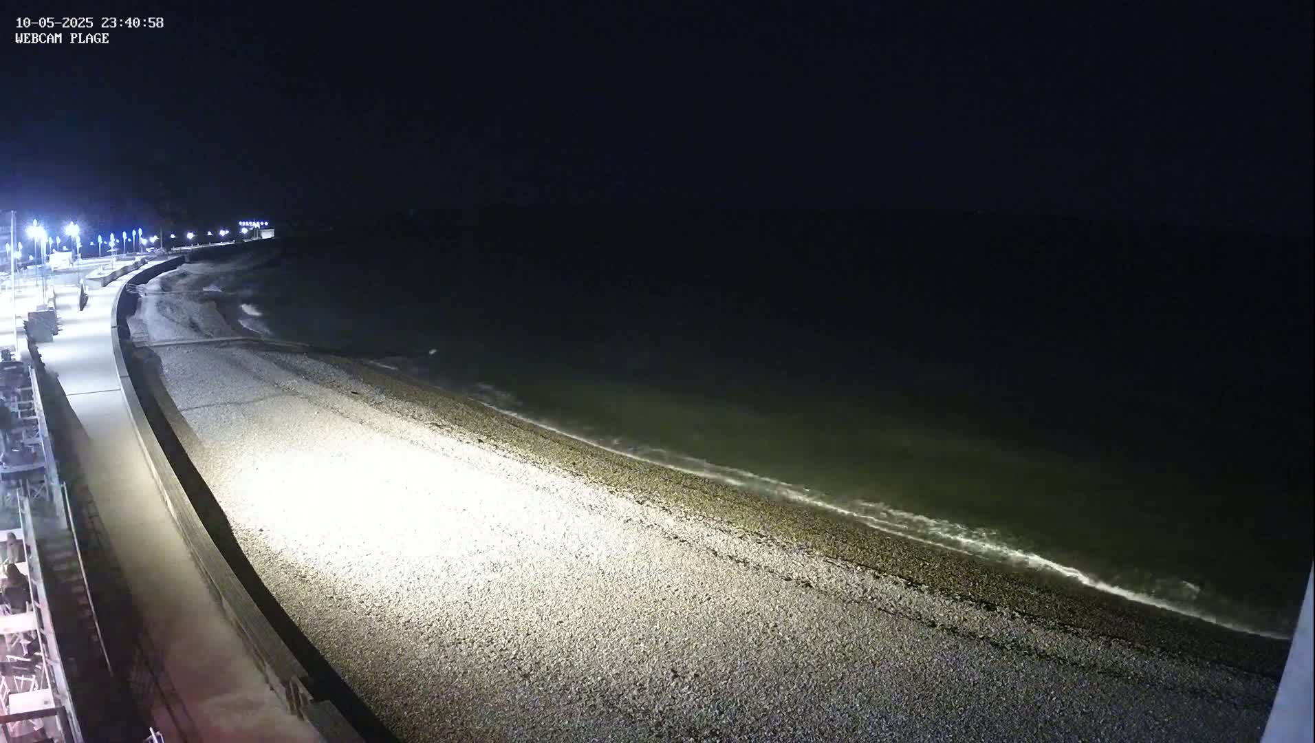 A pebble beach at night is lit by streetlights, with gently lapping waves visible in the dark water.