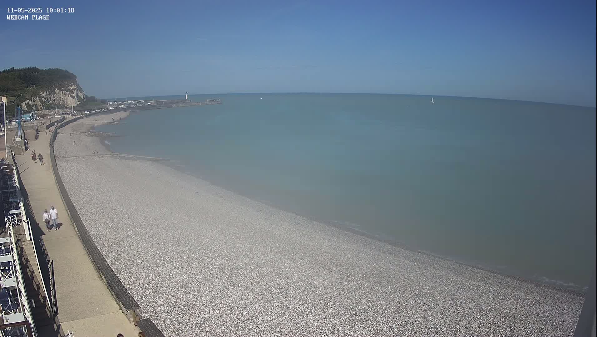 A pebble beach stretches along a calm, light-blue sea under a clear blue sky, with a few people walking along a promenade.