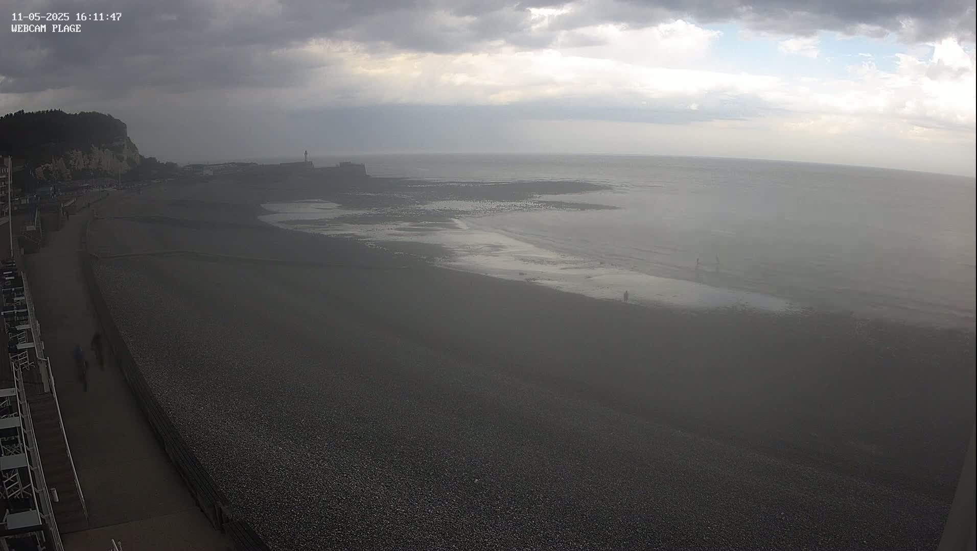A hazy, overcast day reveals a dark pebble beach, a calm sea, and a distant cliffside.