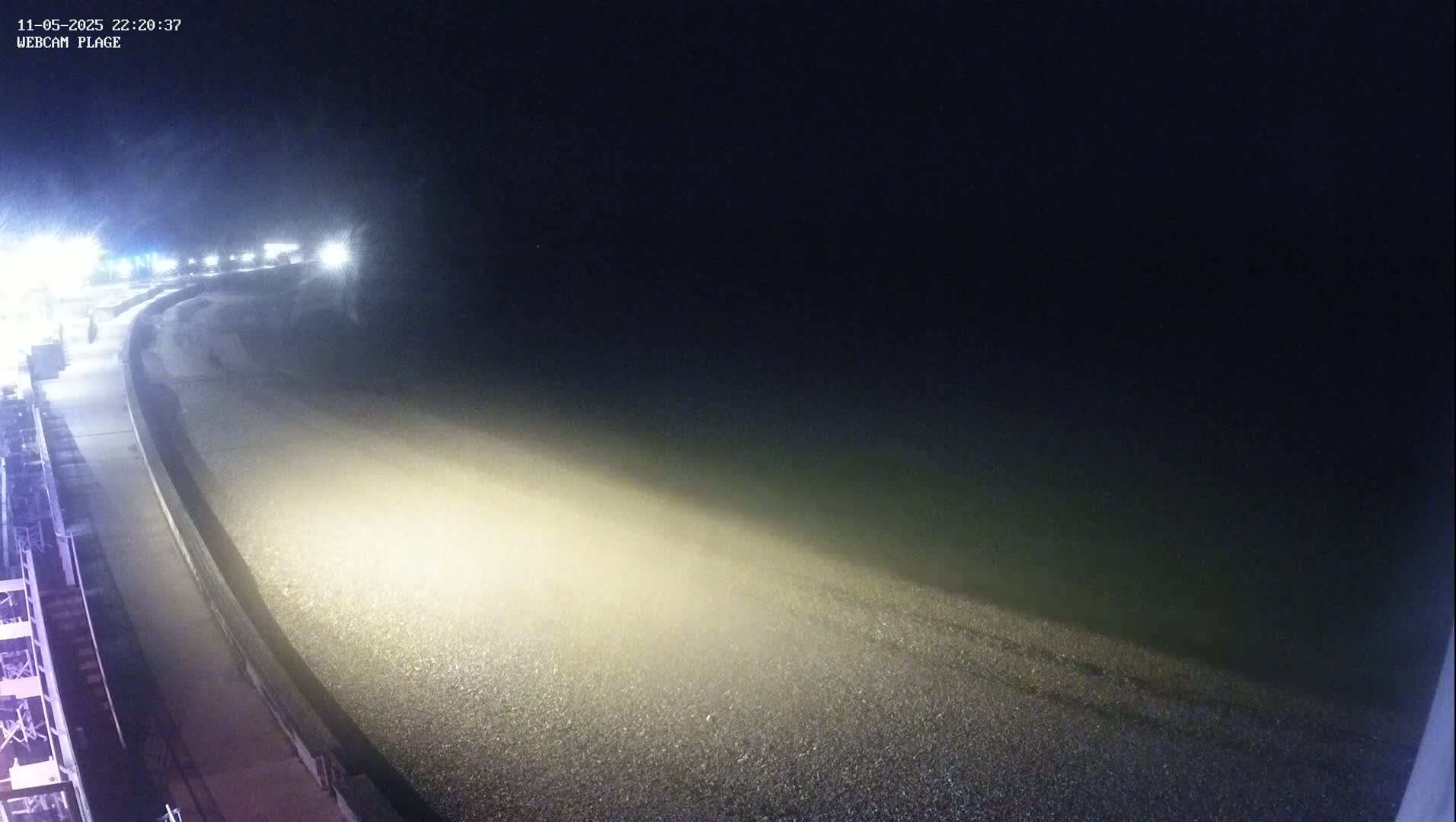 A foggy night scene shows a pebble beach illuminated by a single bright light, with a paved walkway running alongside it.