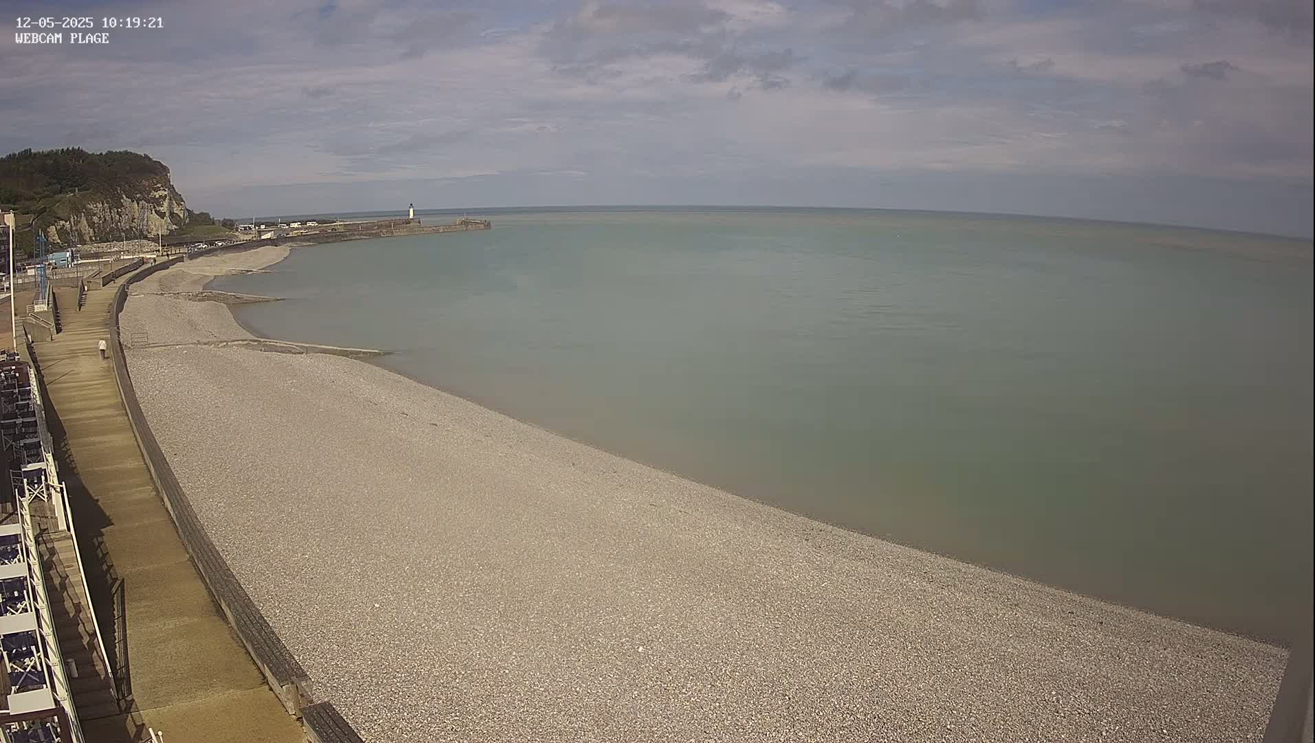 A pebble beach curves gently around a calm, light-green sea, under a mostly cloudy sky.