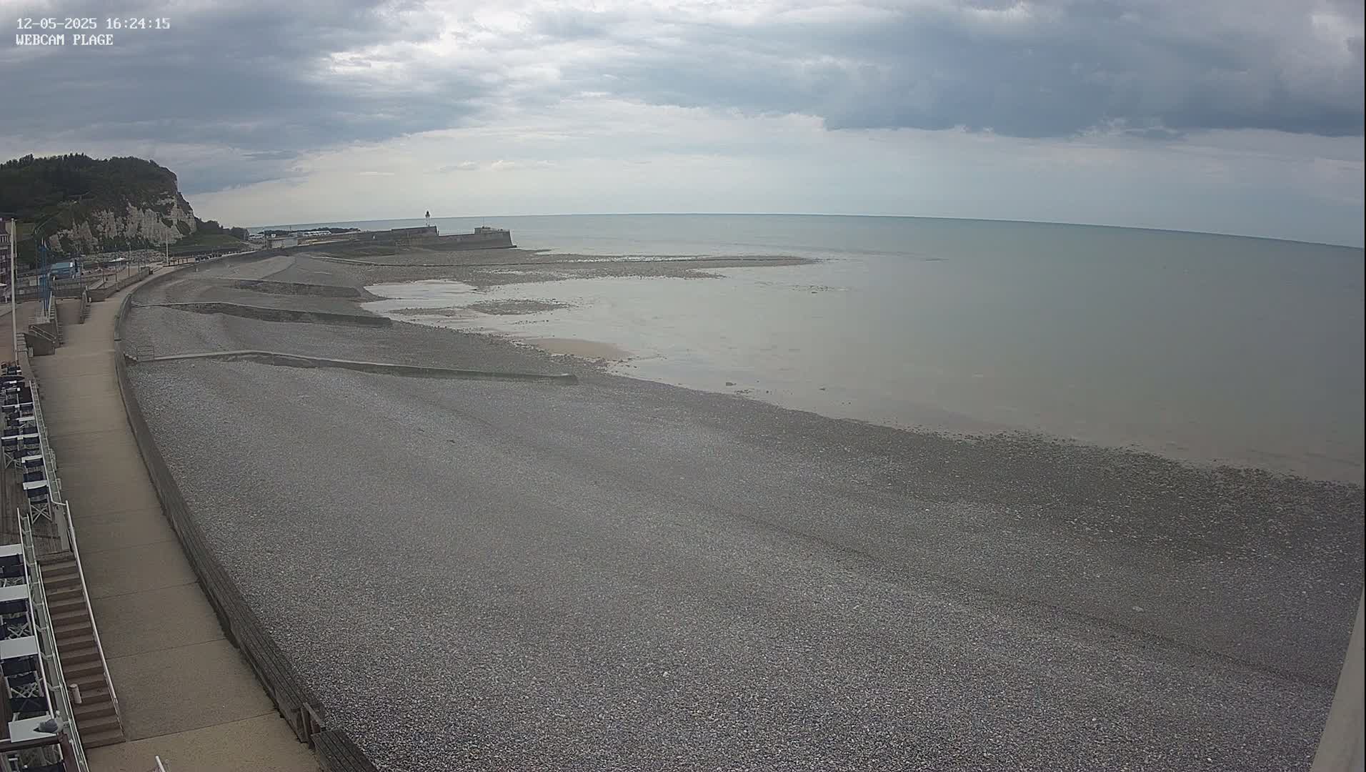 A pebble beach stretches along a paved walkway, under a cloudy sky, toward a calm sea and a distant cliff.