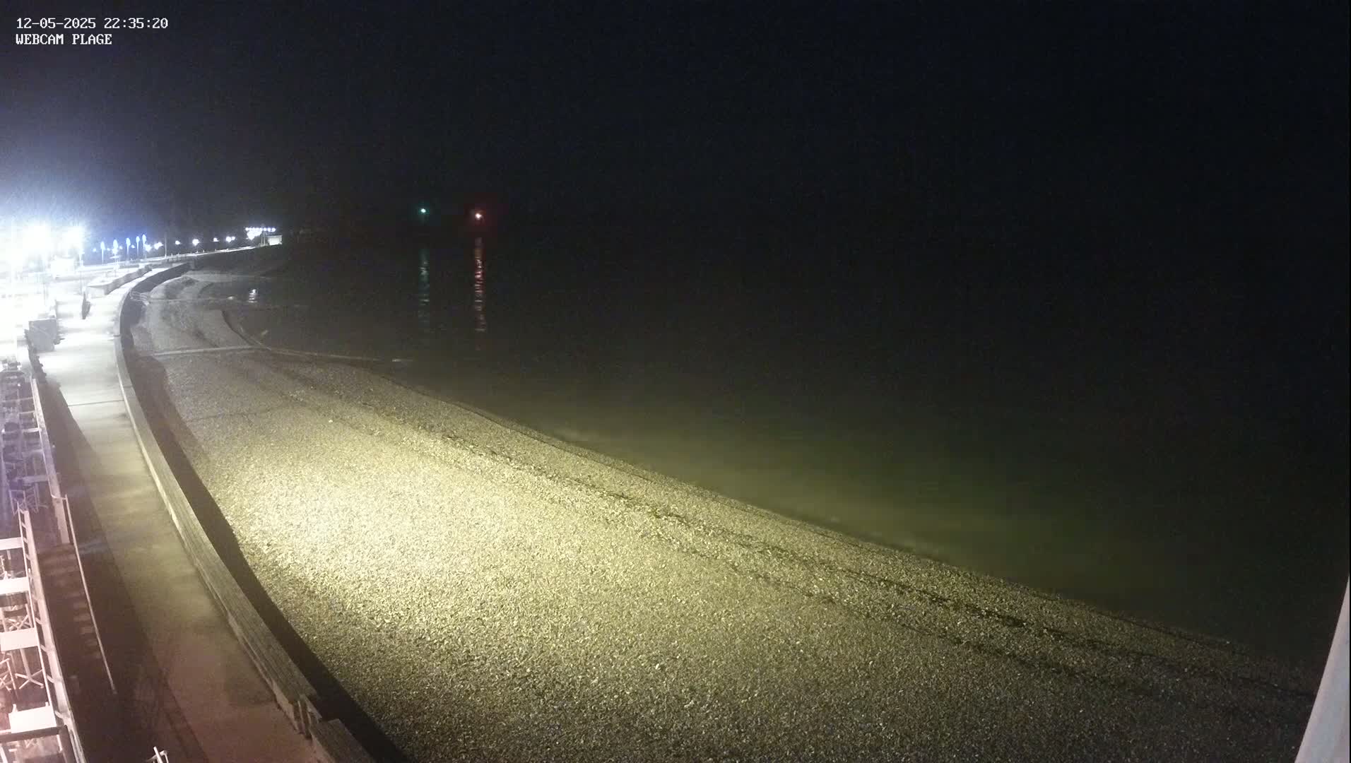 A dark, nighttime view of a pebble beach and a paved walkway beside a calm sea.