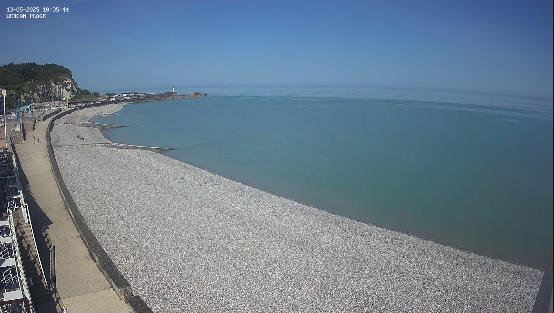 A pebble beach stretches along a calm, teal ocean under a clear blue sky, next to a paved walkway and a cliff in the distance.