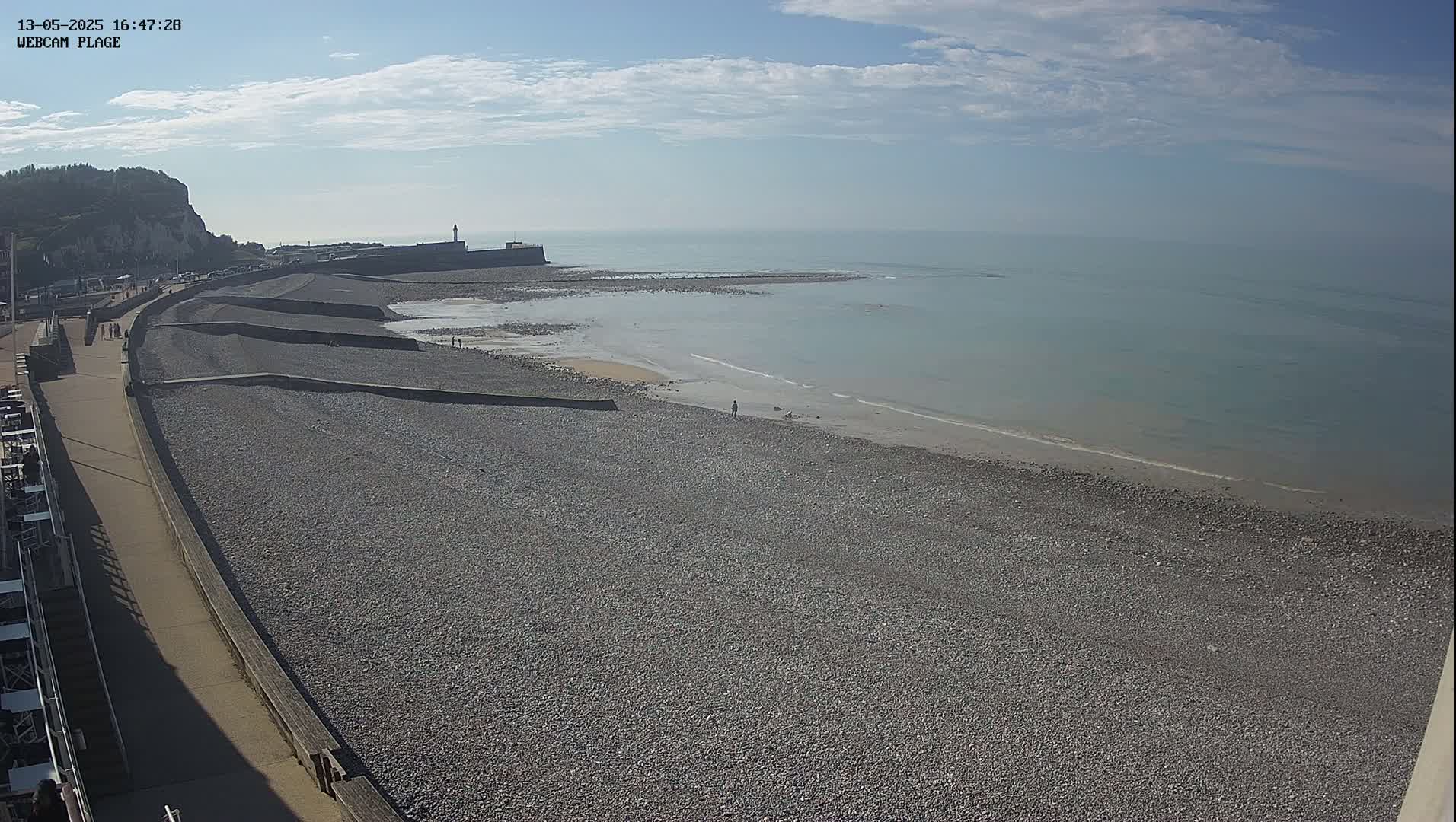 A pebble beach stretches along a calm sea under a mostly clear sky, with a walkway and white cliffs visible in the background.