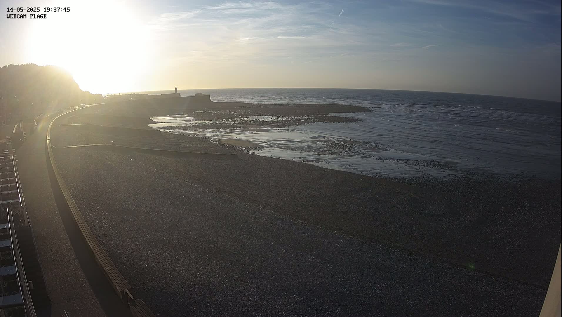 A dark, pebble beach stretches out to a calm, dark-blue sea under a bright, mostly clear sky at sunset.