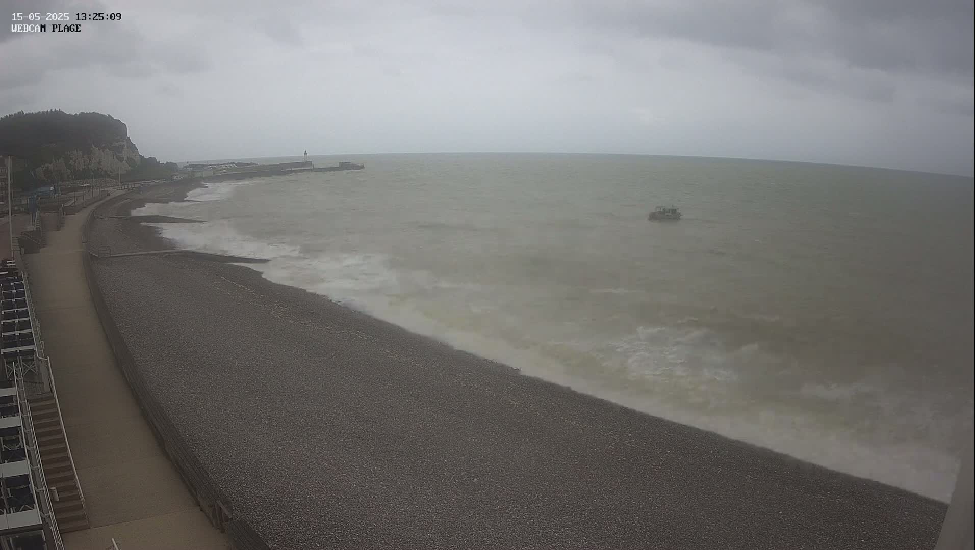A pebble beach with a walkway and a cliff in the background is seen under an overcast sky, with moderate waves crashing on the shore.