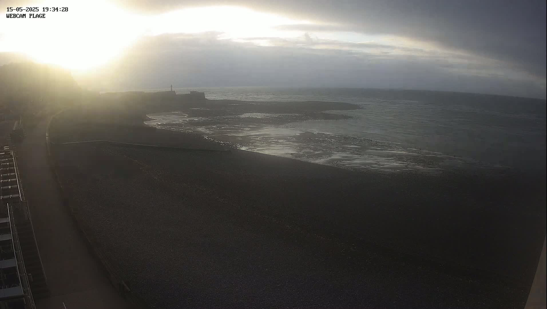 A dark, pebbly beach stretches out to a calm sea under an overcast sky, with the sun shining brightly through the clouds near the horizon.