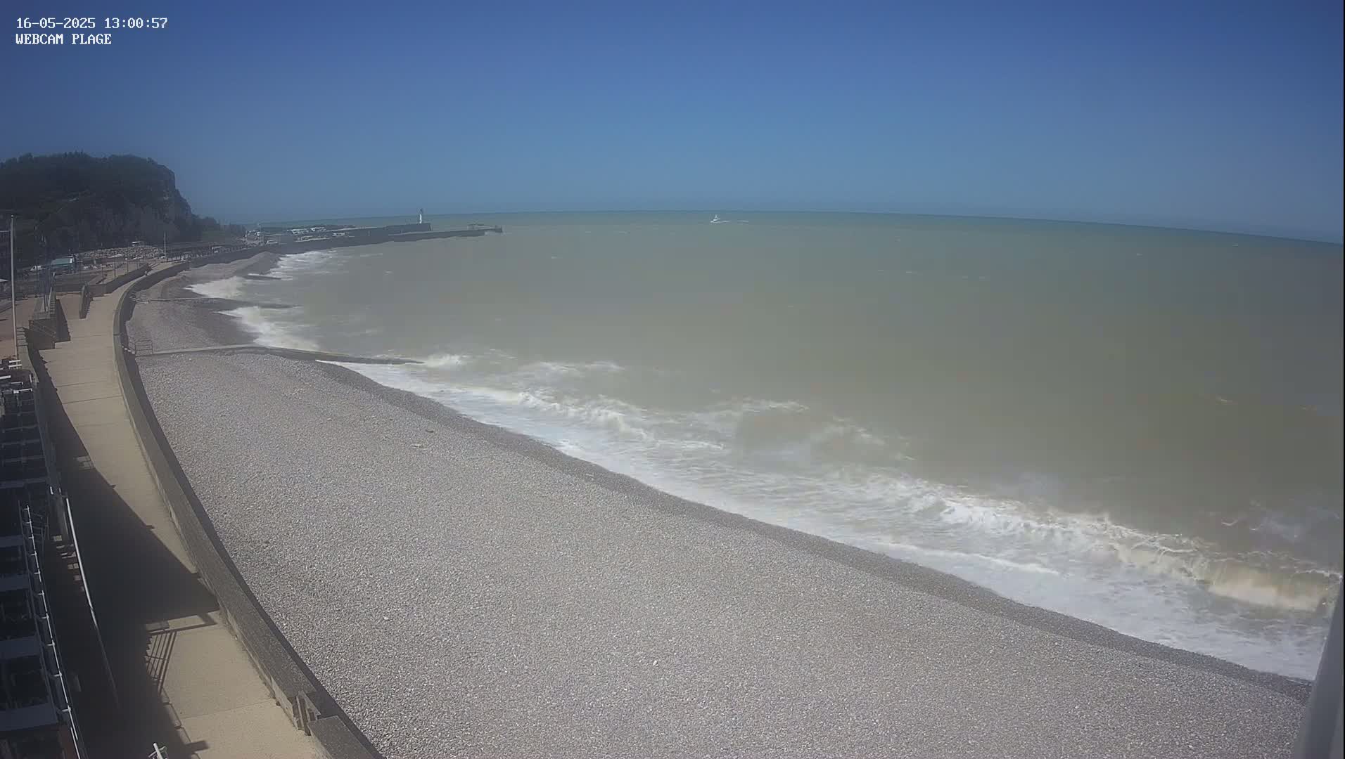 A pebble beach with gentle waves rolling onto the shore on a clear, sunny day, next to a paved walkway and a cliff in the distance.
