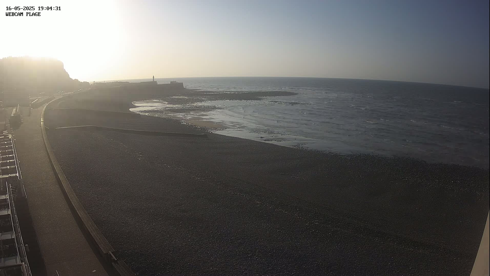 A hazy sunrise illuminates a dark pebble beach and calm ocean, with a paved walkway and distant buildings visible.