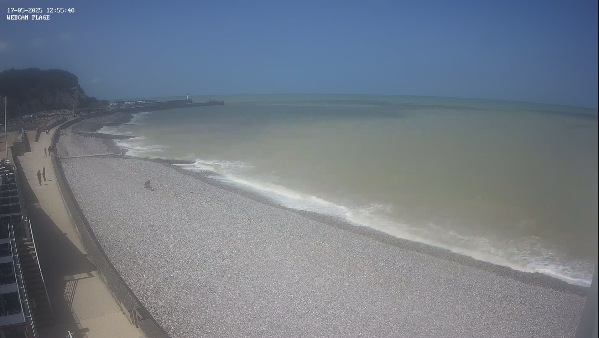 A pebble beach with a few people walking along a paved path next to it, under a clear blue sky, with gentle waves rolling onto the shore.