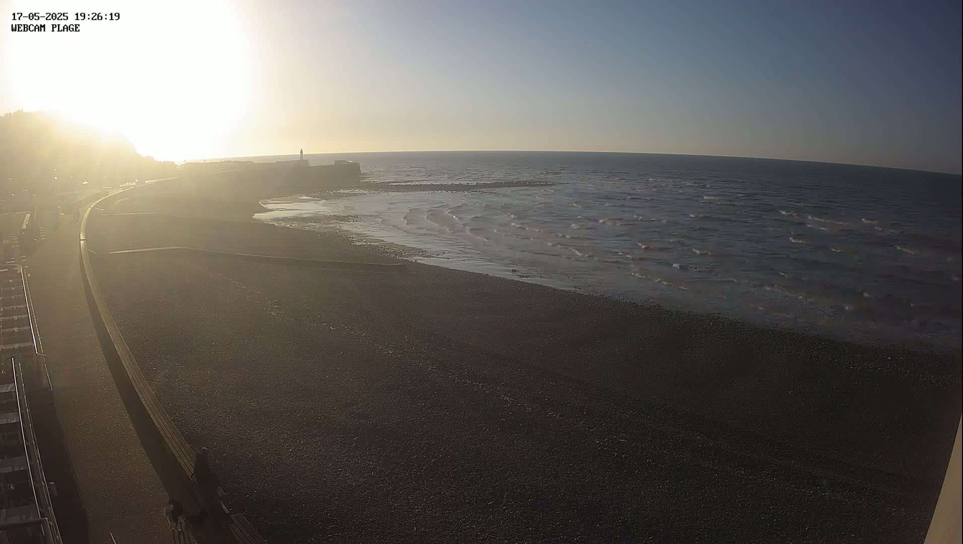 A dark, pebble beach meets a calm, dark-blue ocean under a bright, hazy sunrise.
