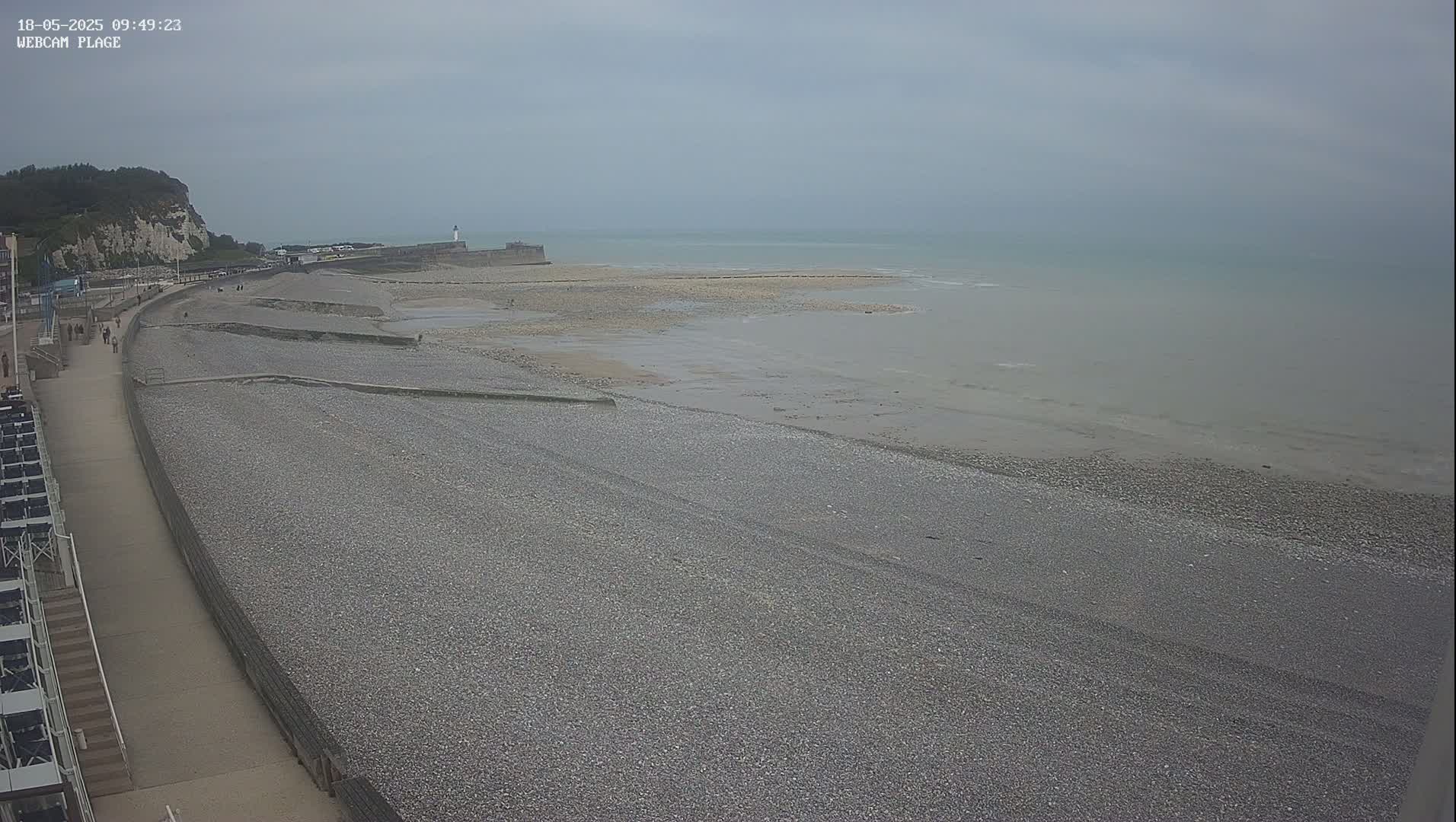 A pebbled beach stretches along a calm, overcast ocean, with a walkway and white cliffs visible in the background.
