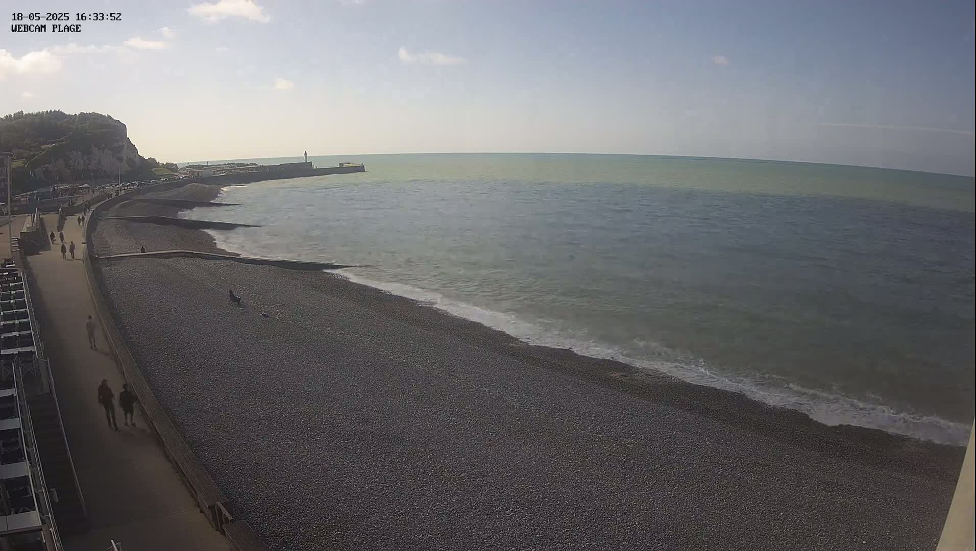 A pebble beach with a walkway and people on it is next to a calm sea under a mostly sunny sky.