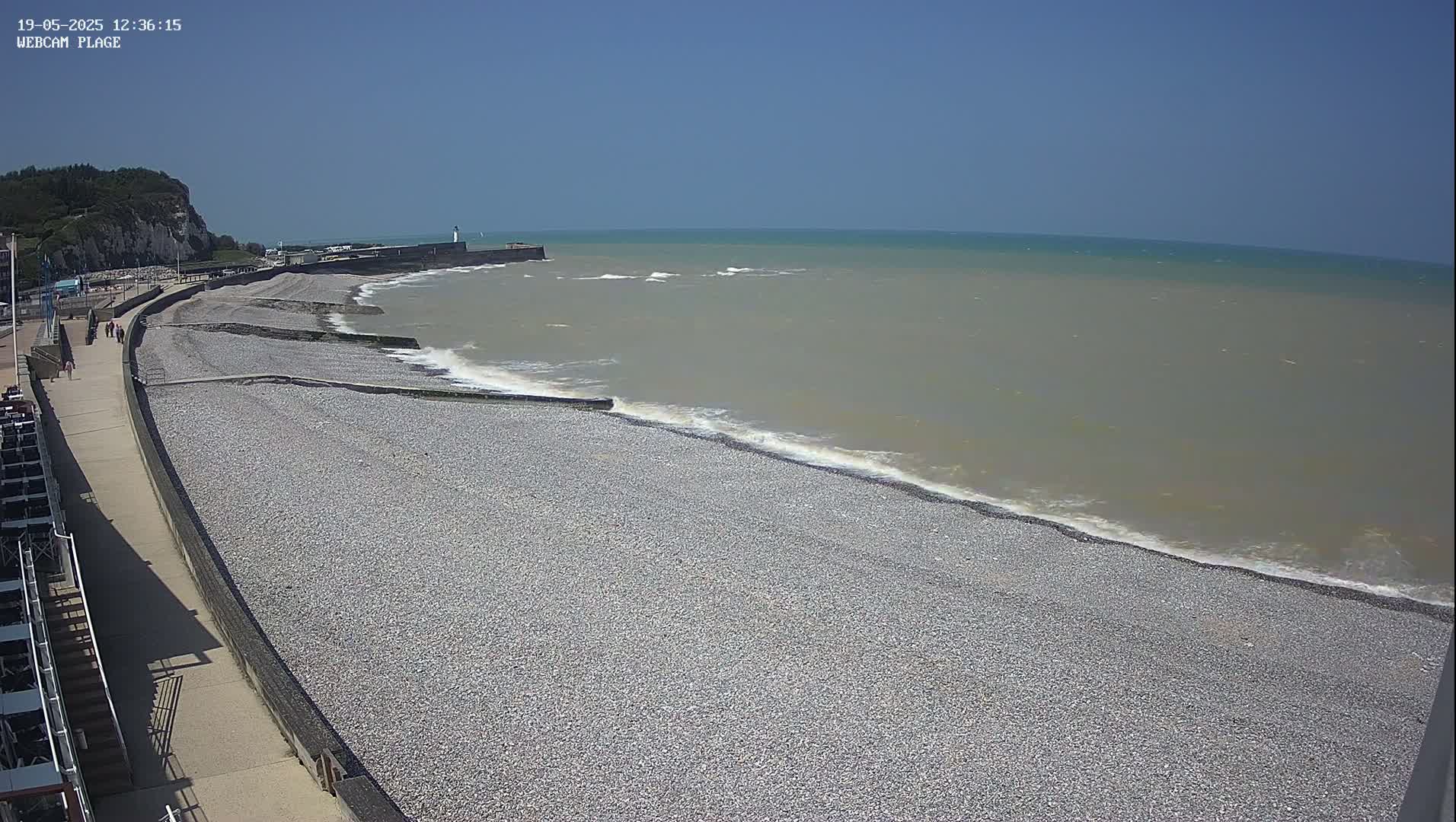 A pebble beach stretches along a calm, grayish-green sea under a clear blue sky, with a paved walkway and a few people visible in the distance.