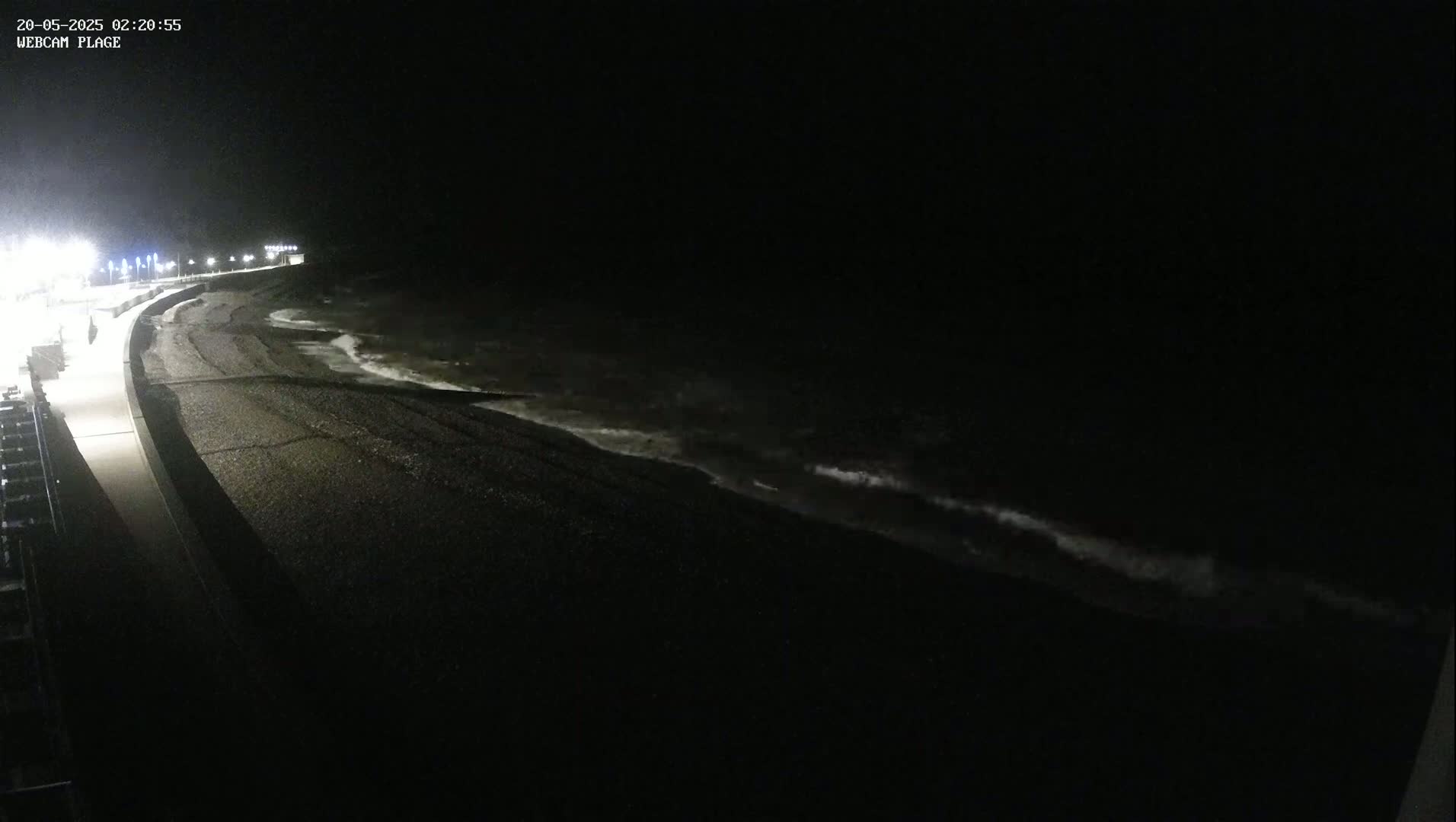 A dark, nighttime beach scene shows a dimly lit paved walkway next to a dark, pebble-covered shore where small waves are lapping at the edge of the water under a black sky.