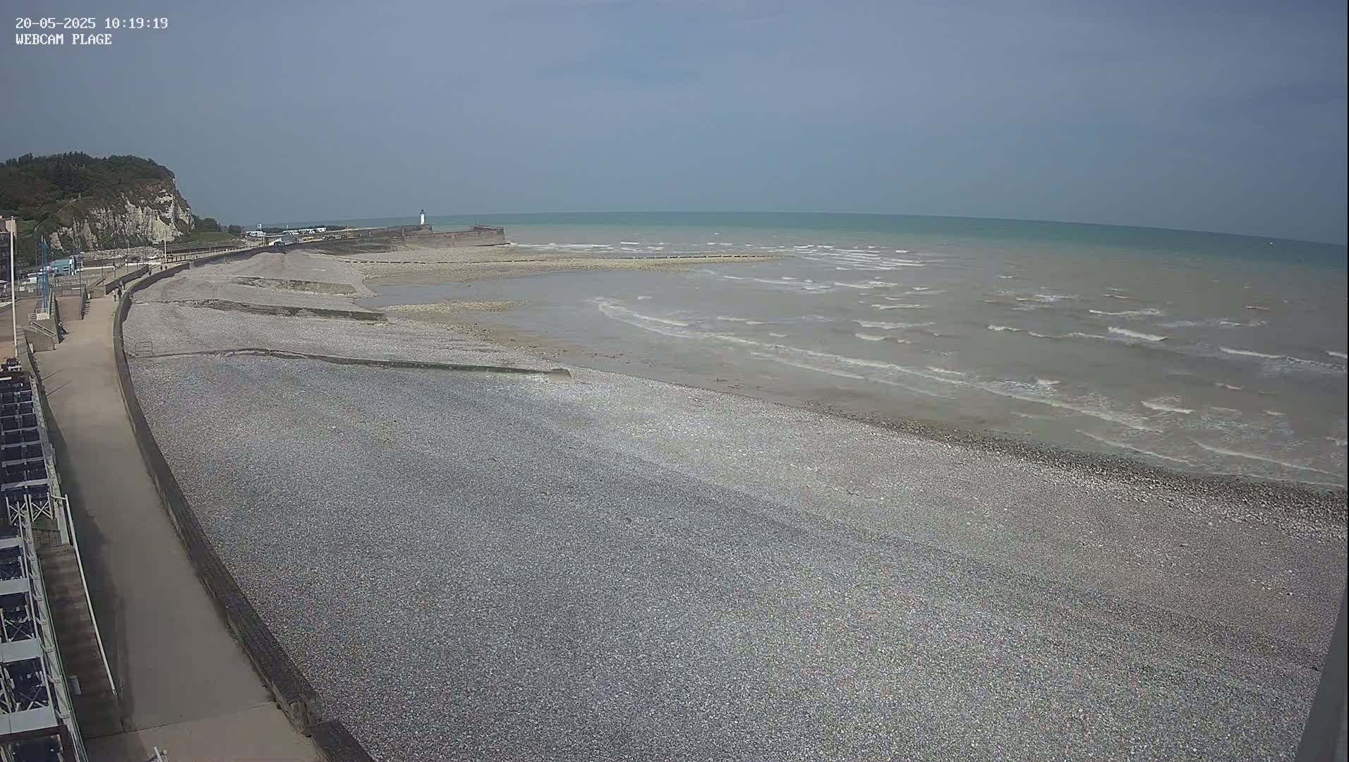 A pebble beach stretches along a calm sea under a clear sky, with a paved walkway and a cliff visible in the background.