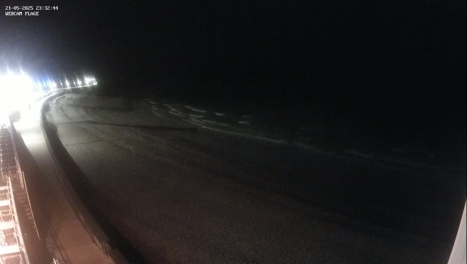A dark, nighttime beach scene shows gently lapping waves on a mostly empty shore, with a paved walkway curving along the upper left.