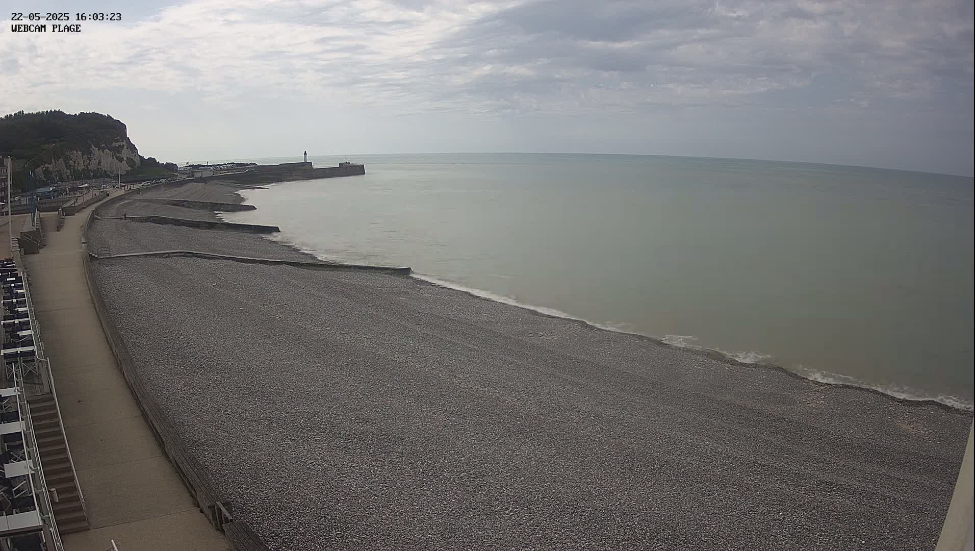 A pebble beach stretches along a calm, gray sea under an overcast sky, with a walkway and buildings visible in the background.