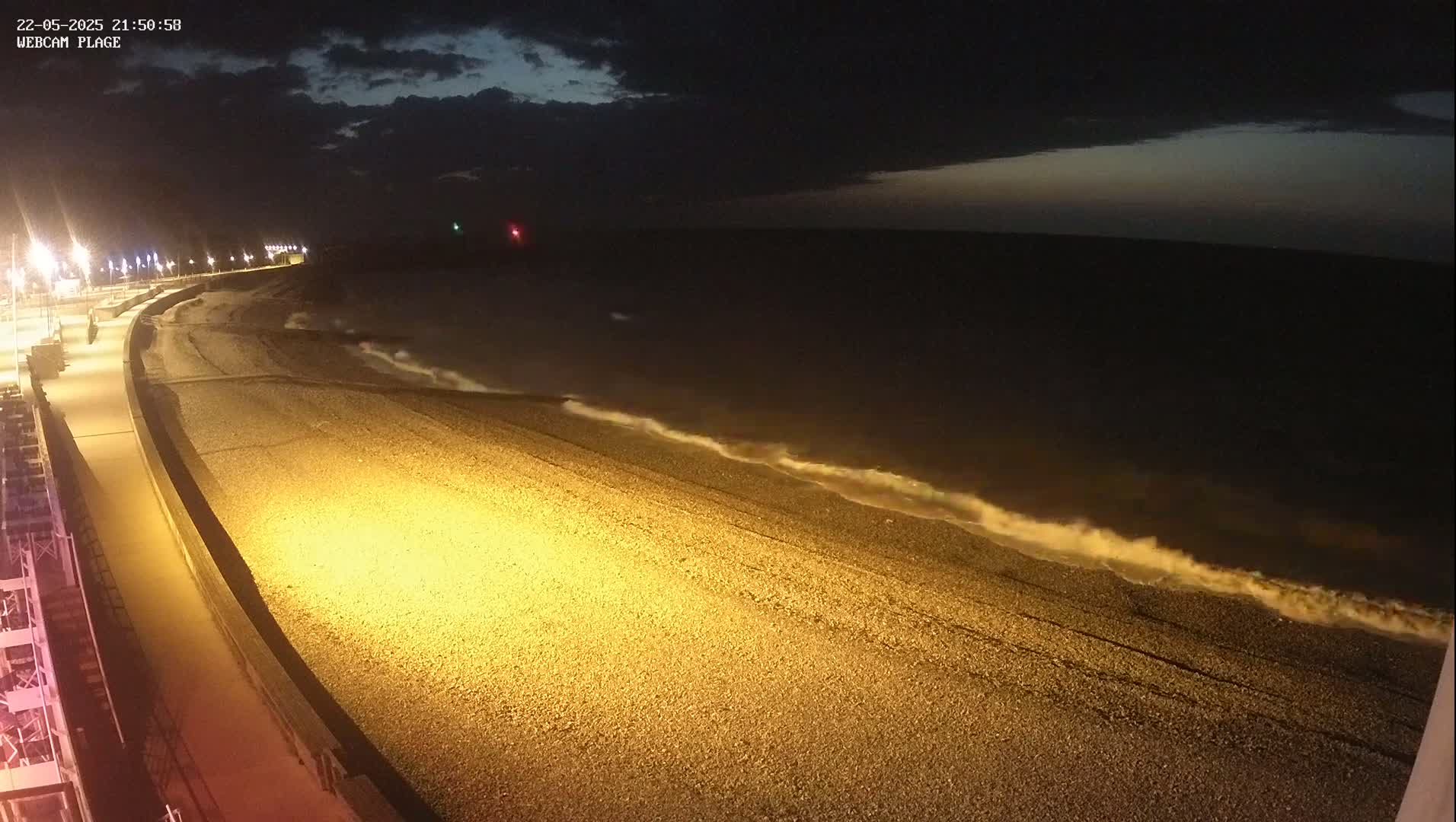 A dark, nighttime view of a pebble beach with gentle waves lapping the shore, illuminated by artificial lights along a paved walkway.