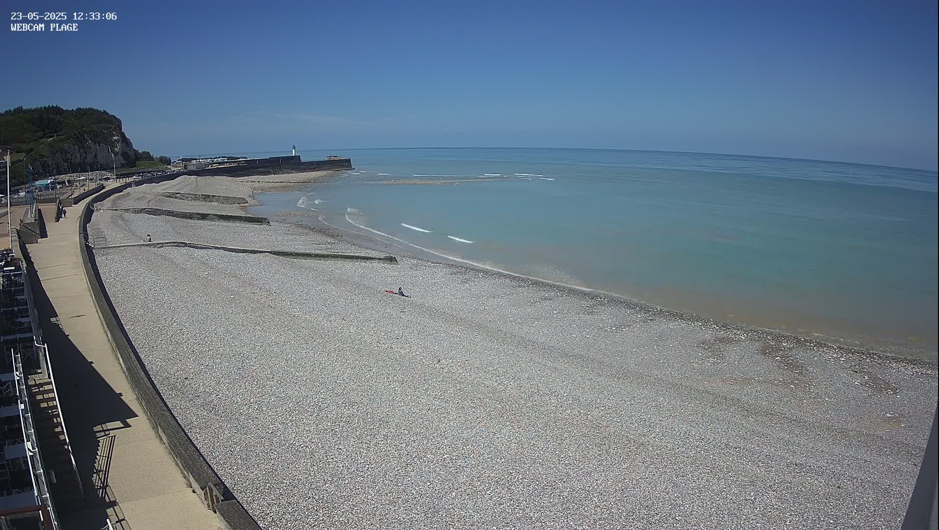 A pebble beach stretches along a calm, light-blue sea under a clear blue sky, with a walkway and a few people visible.