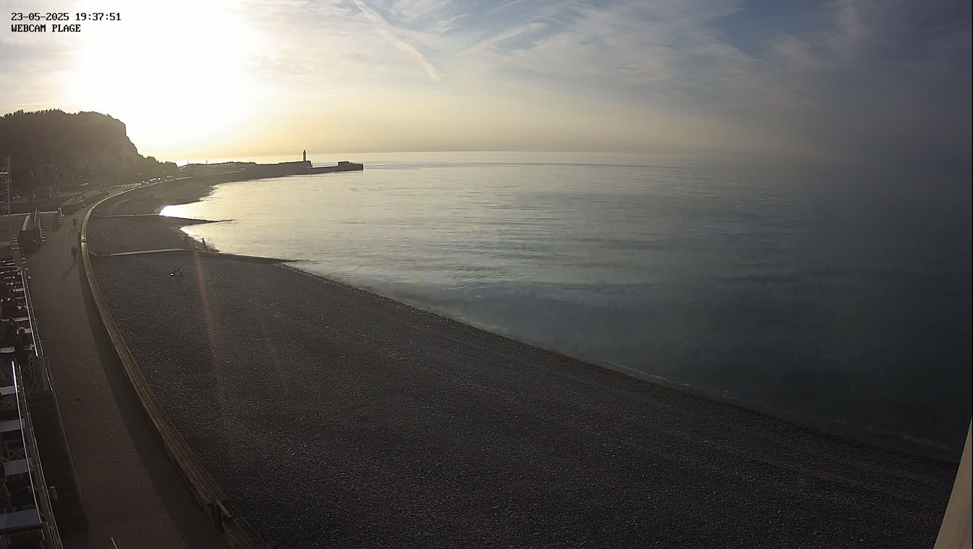 A dark pebble beach curves gently around a calm sea under a mostly clear, pale sky at sunset, with a small pier visible in the distance.
