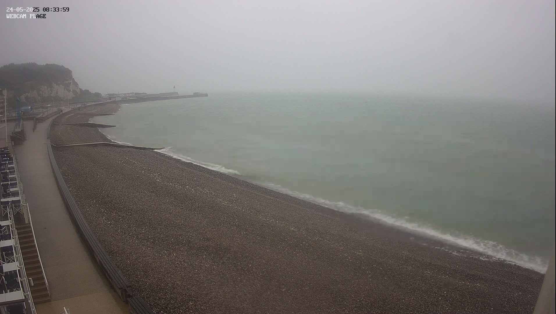 A rocky beach and calm, grey sea are visible under an overcast sky on a misty or rainy day.