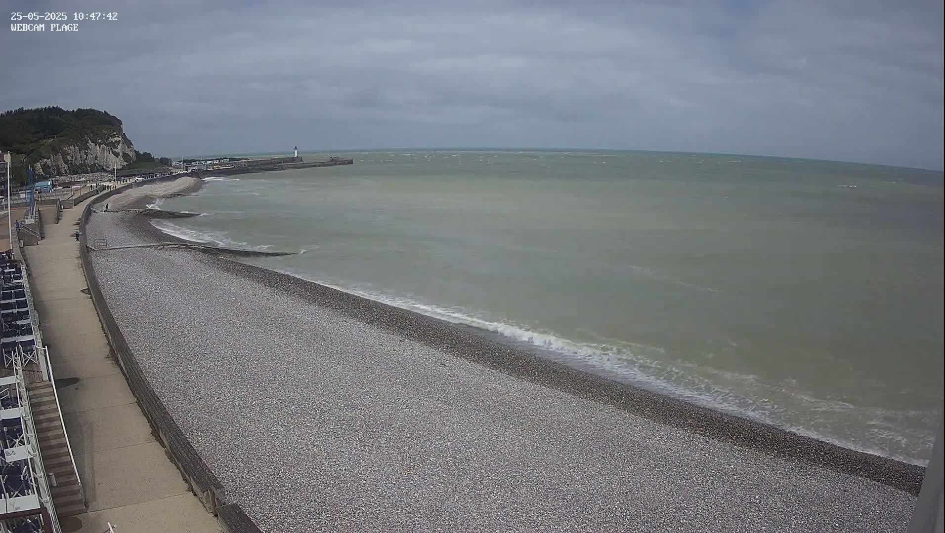 A pebble beach and a paved walkway stretch alongside a calm, gray sea under an overcast sky.