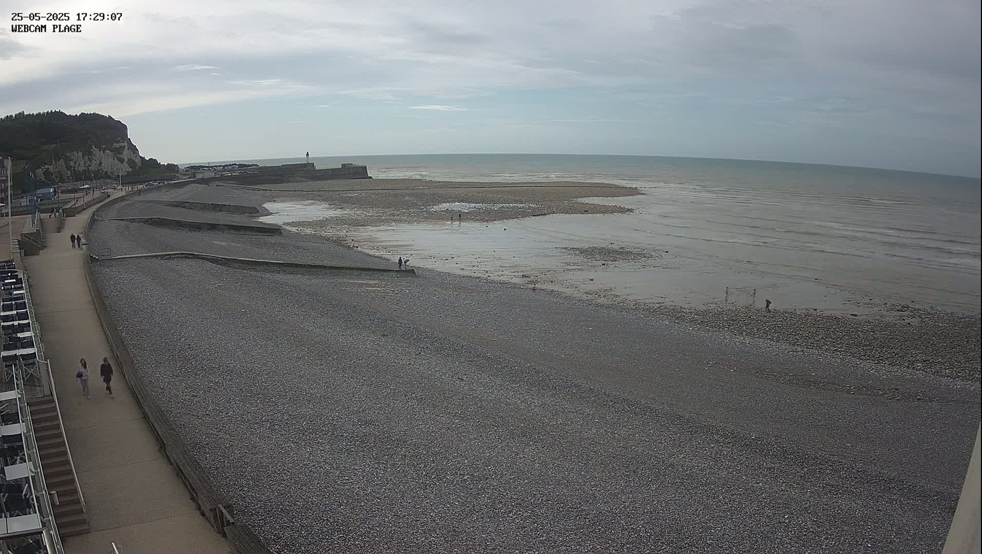 A pebble beach stretches out to a calm, gray sea under an overcast sky, with a few people walking on the beach and along a nearby promenade.