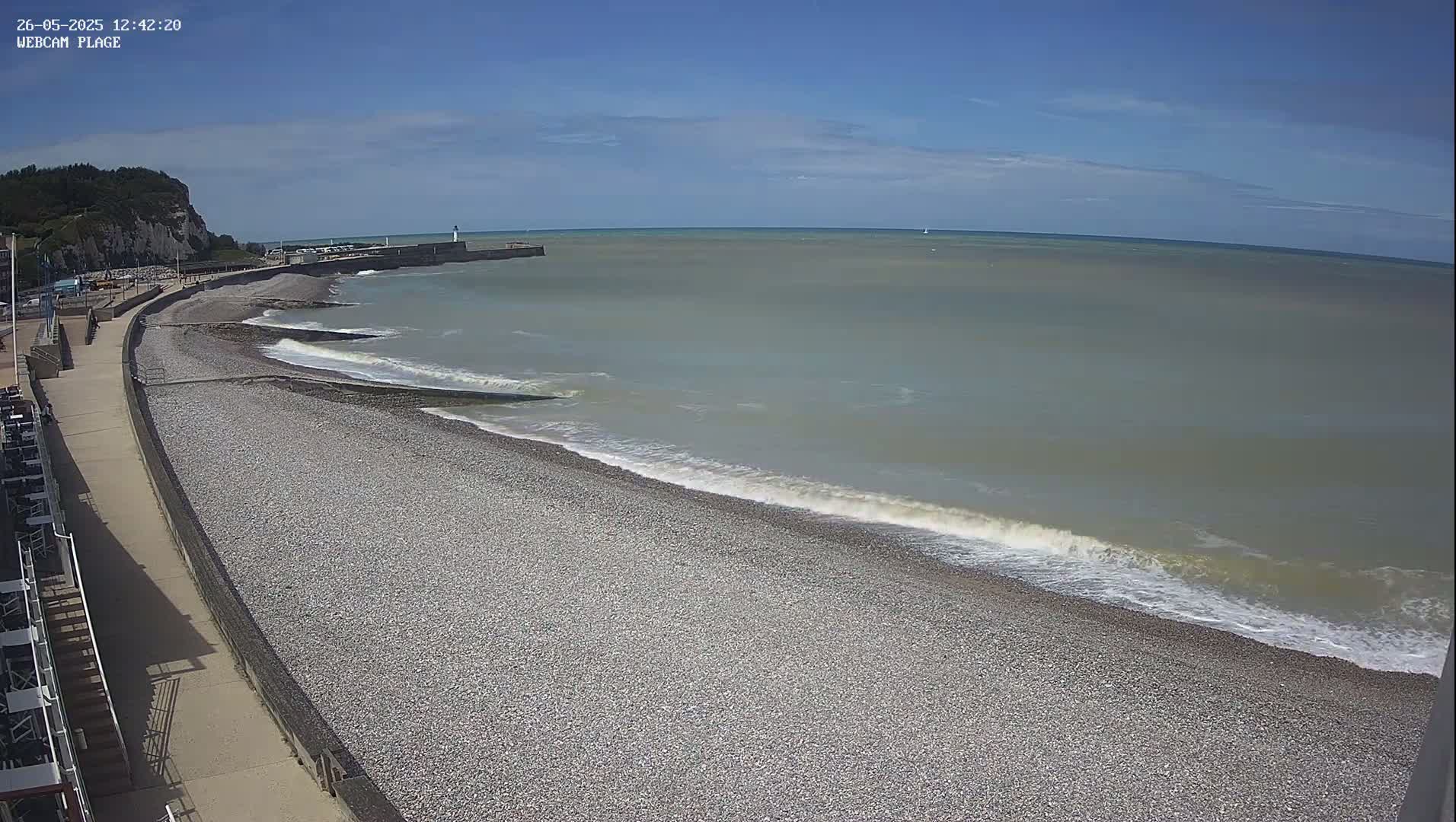 A pebble beach with gentle waves rolls onto the shore under a clear, sunny sky, next to a paved walkway and a cliff in the distance.