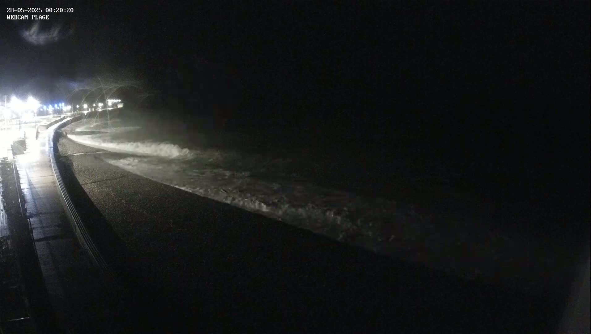 At night, waves crash against a dark, pebbled shoreline next to a paved walkway lit by streetlights.