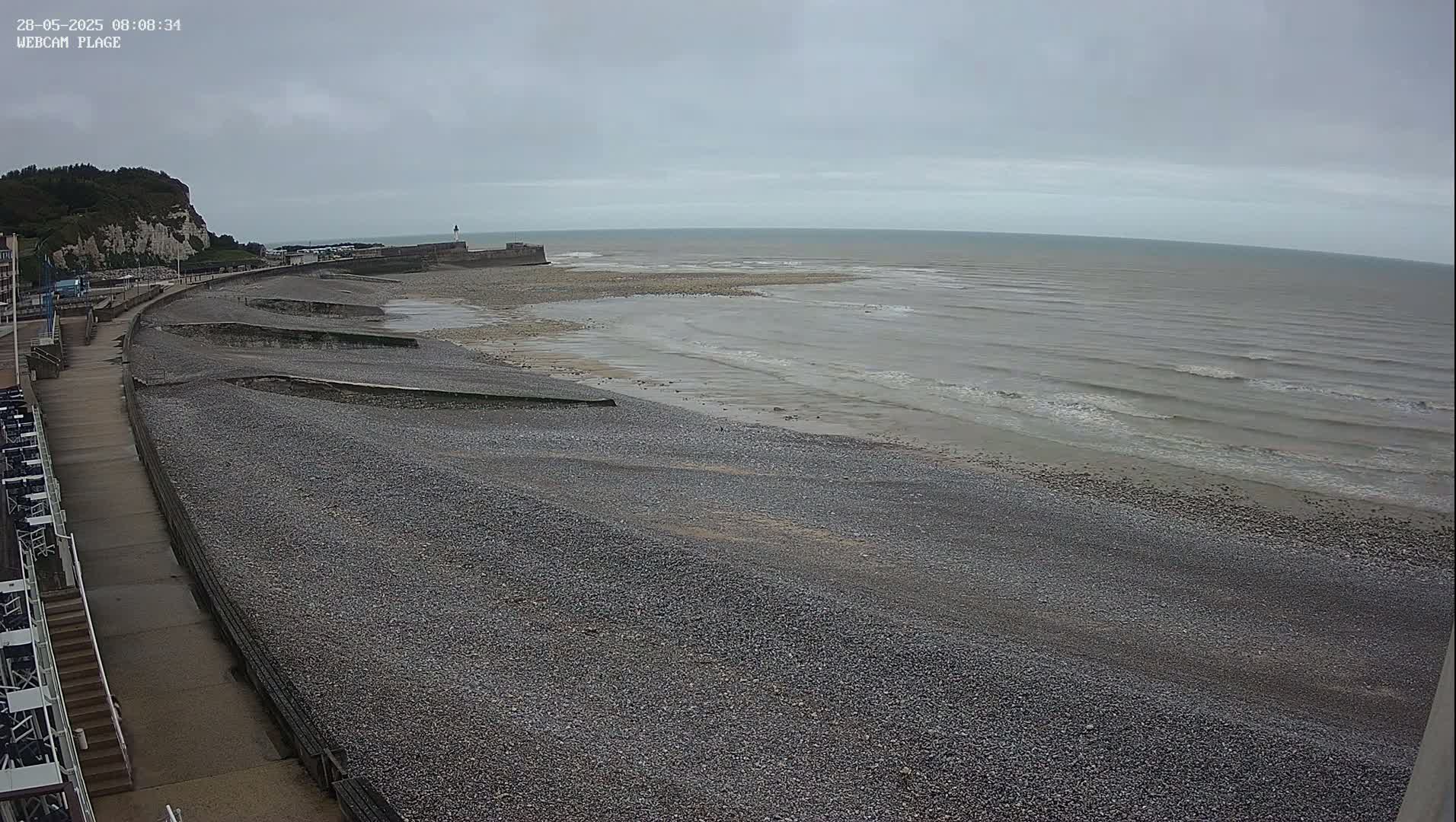 A pebble beach stretches along a coastline under an overcast sky, with gentle waves lapping the shore near a low-lying seawall and a cliff in the background.