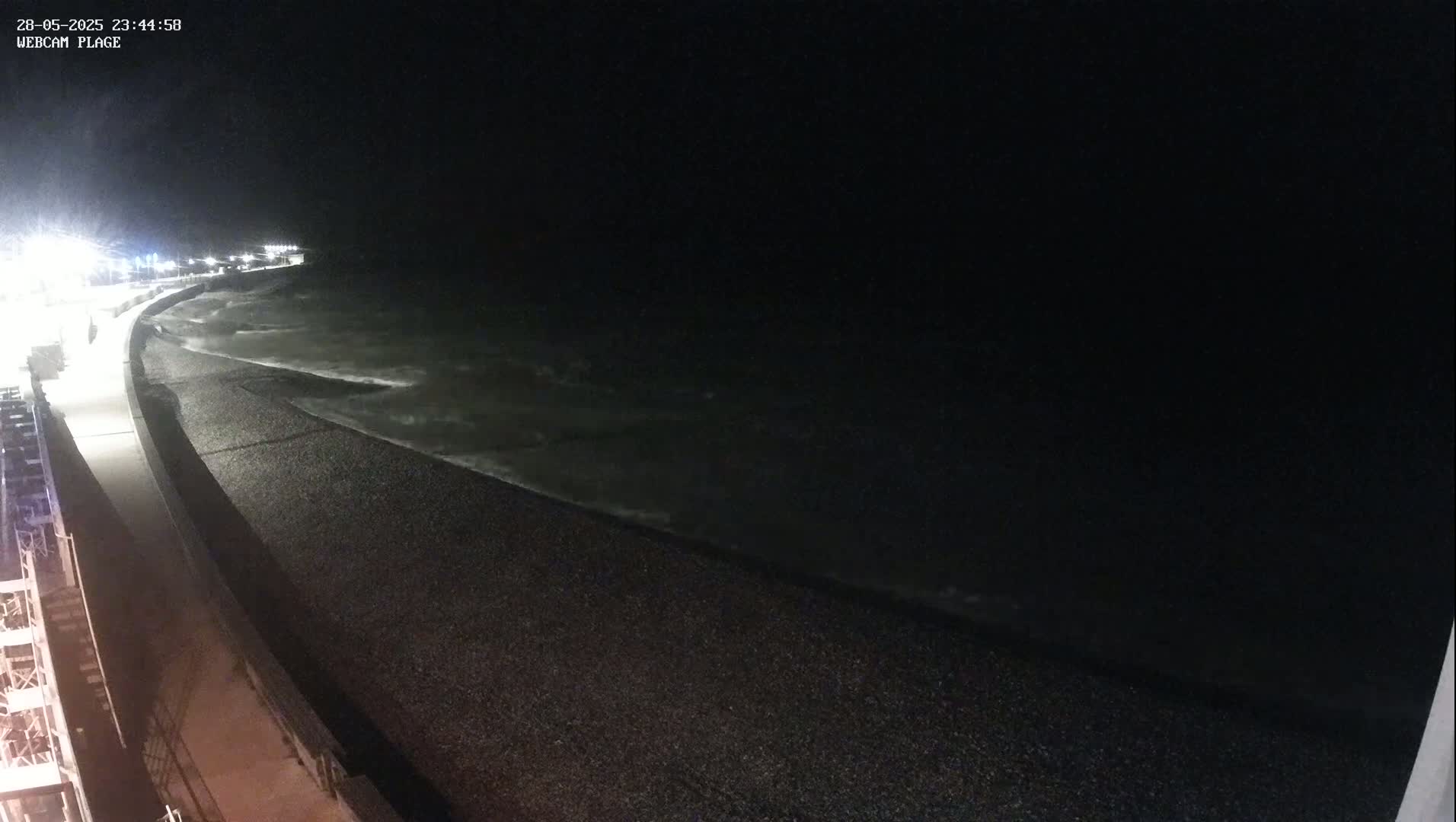 A dark, nighttime beach scene shows a pebble shoreline with slightly wavy water under a clear, starless sky.